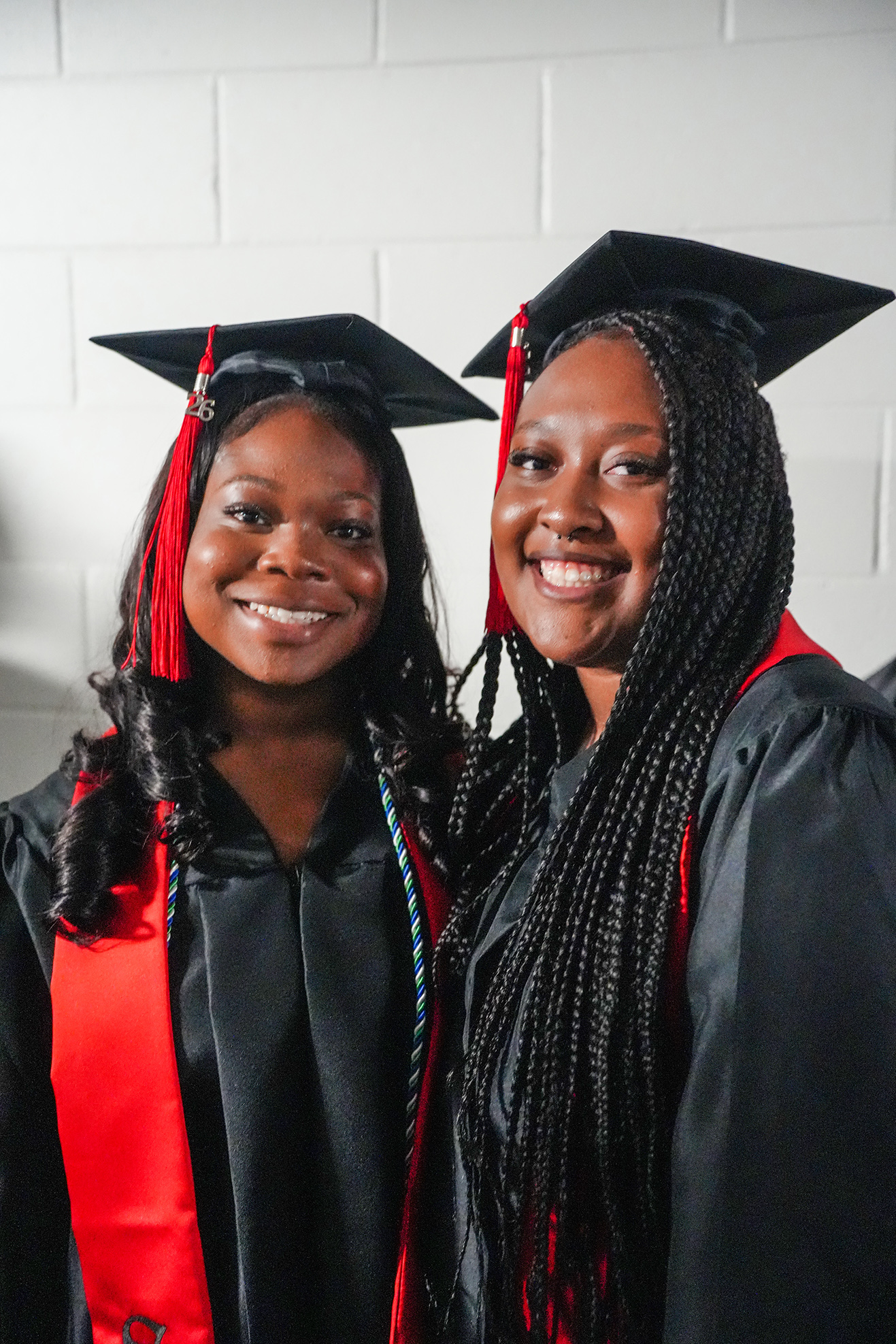 Two women in black graduation gowns with red stoles and black caps with tassels smile and stand close together.