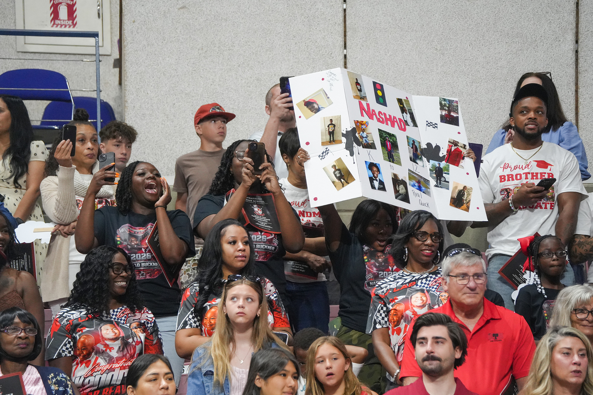 Spectators at a sports event, one holding a poster with images and text, others holding up phones and clapping.