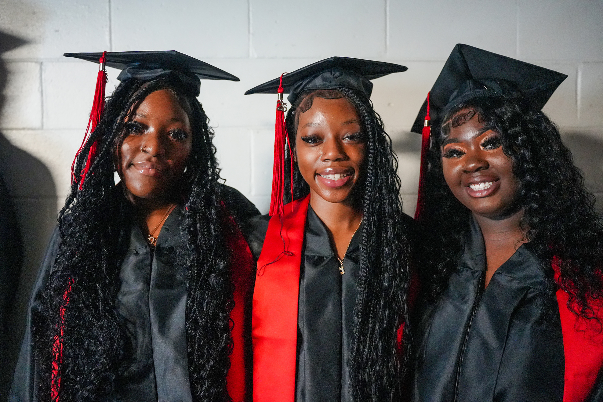 Three women in caps and gowns pose for a photo. They have long, curly hair and wear black and red graduation attire.