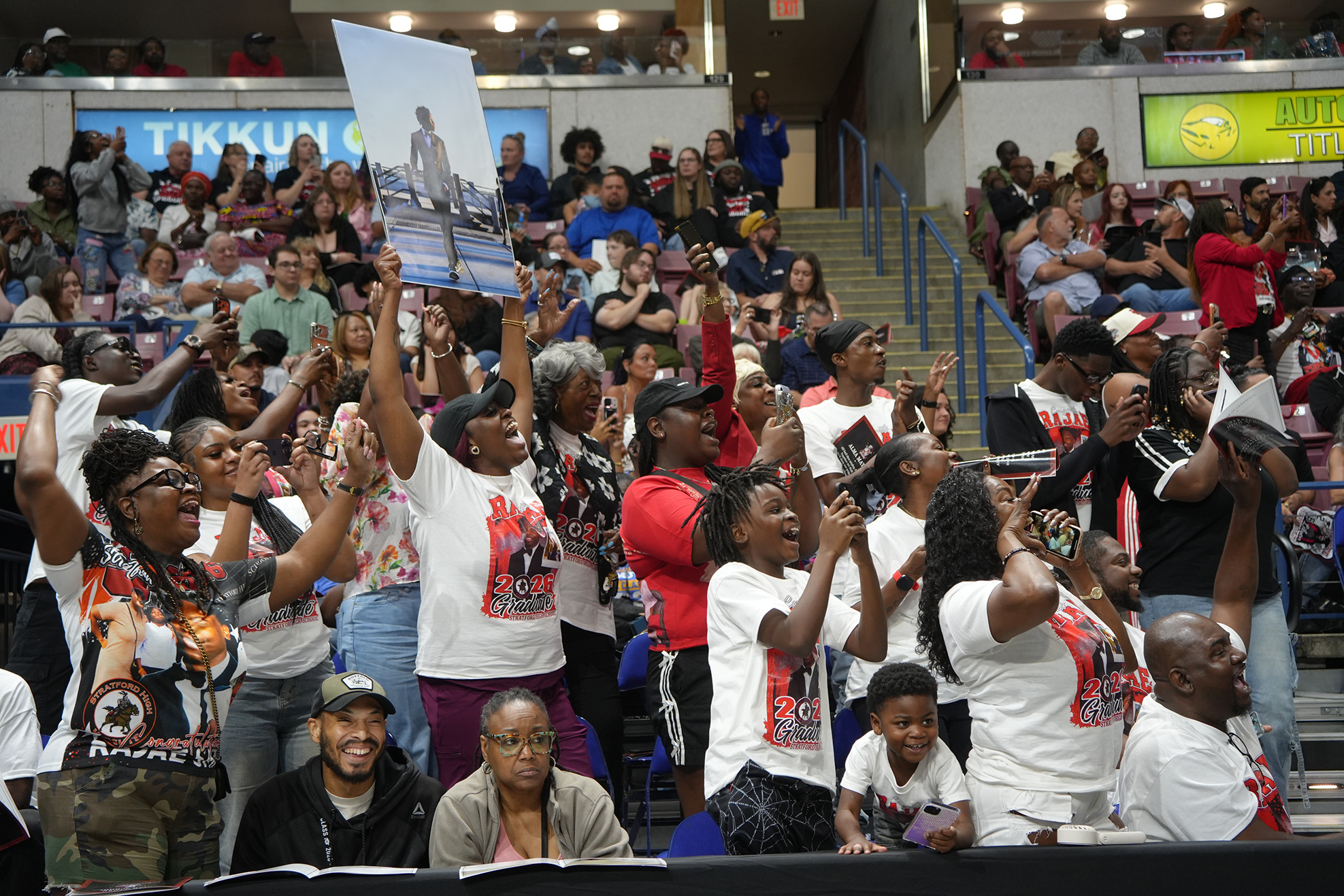 Large group of people in a stadium, cheering and holding up signs, with banners and advertisements visible.