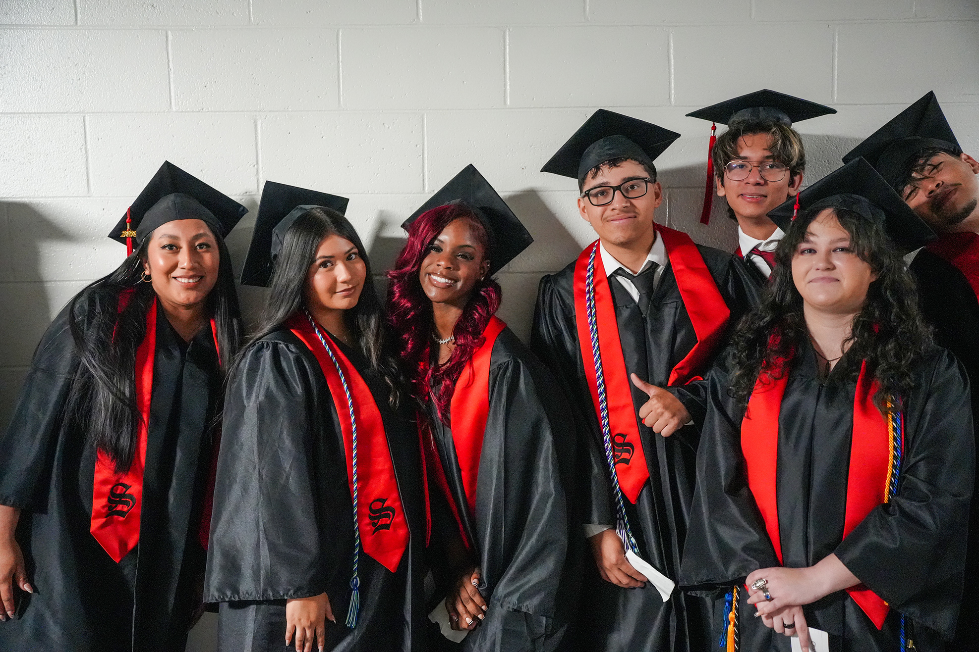 Graduates in black and red robes and caps with tassels pose together for a photo against a white wall.