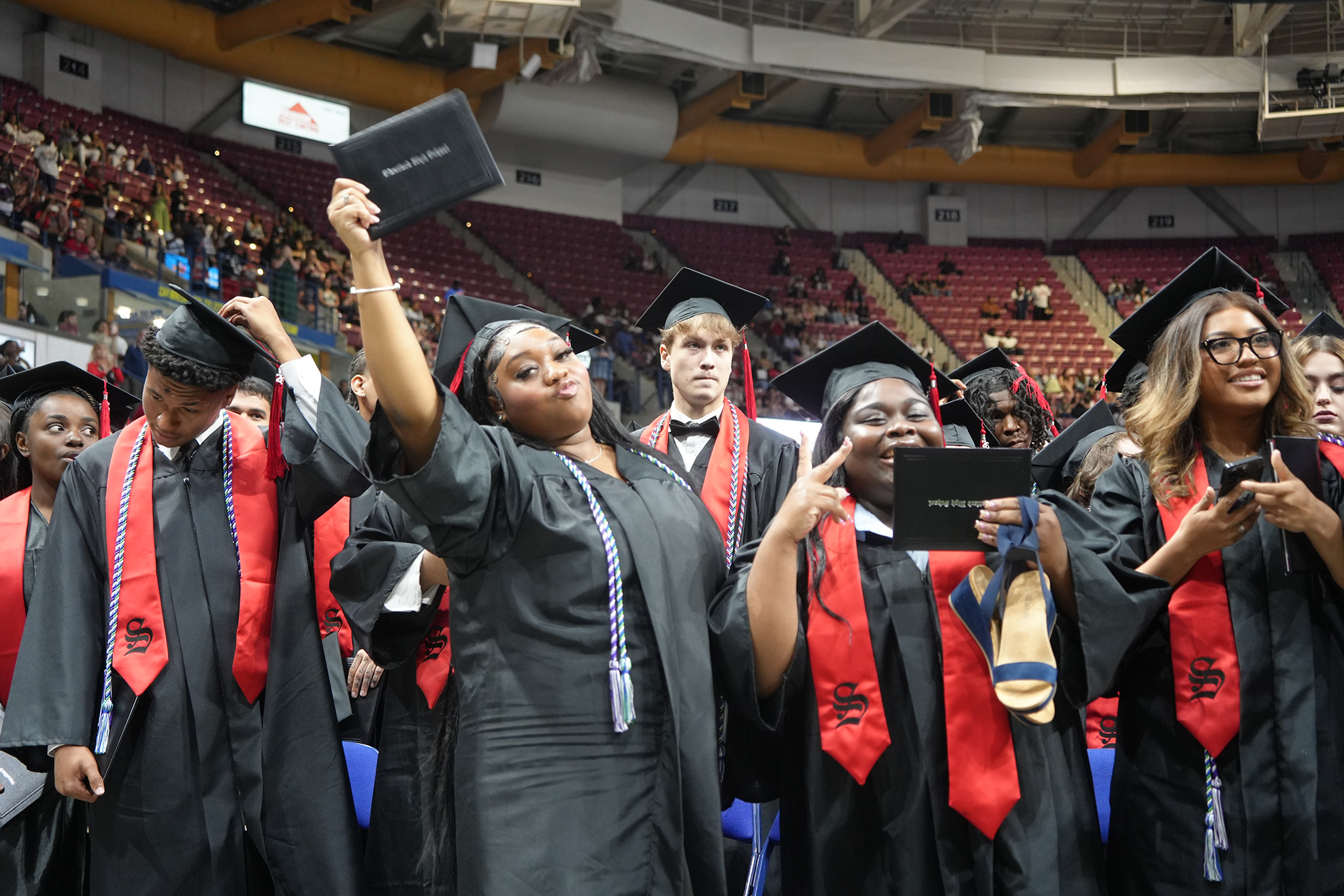 A group of people in black graduation gowns and red stoles, raising diplomas in an indoor stadium.