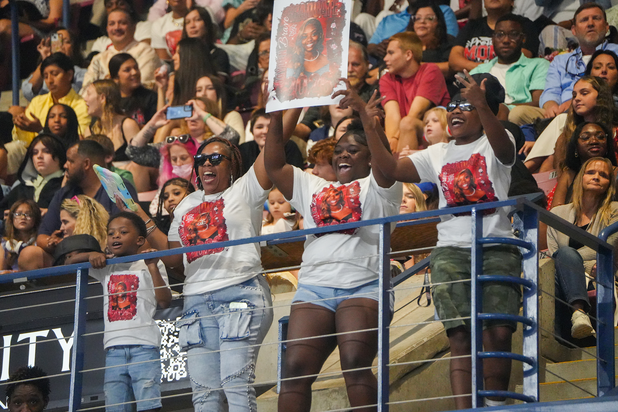 A group of people in matching shirts standing on a balcony, holding up a photo in a stadium.