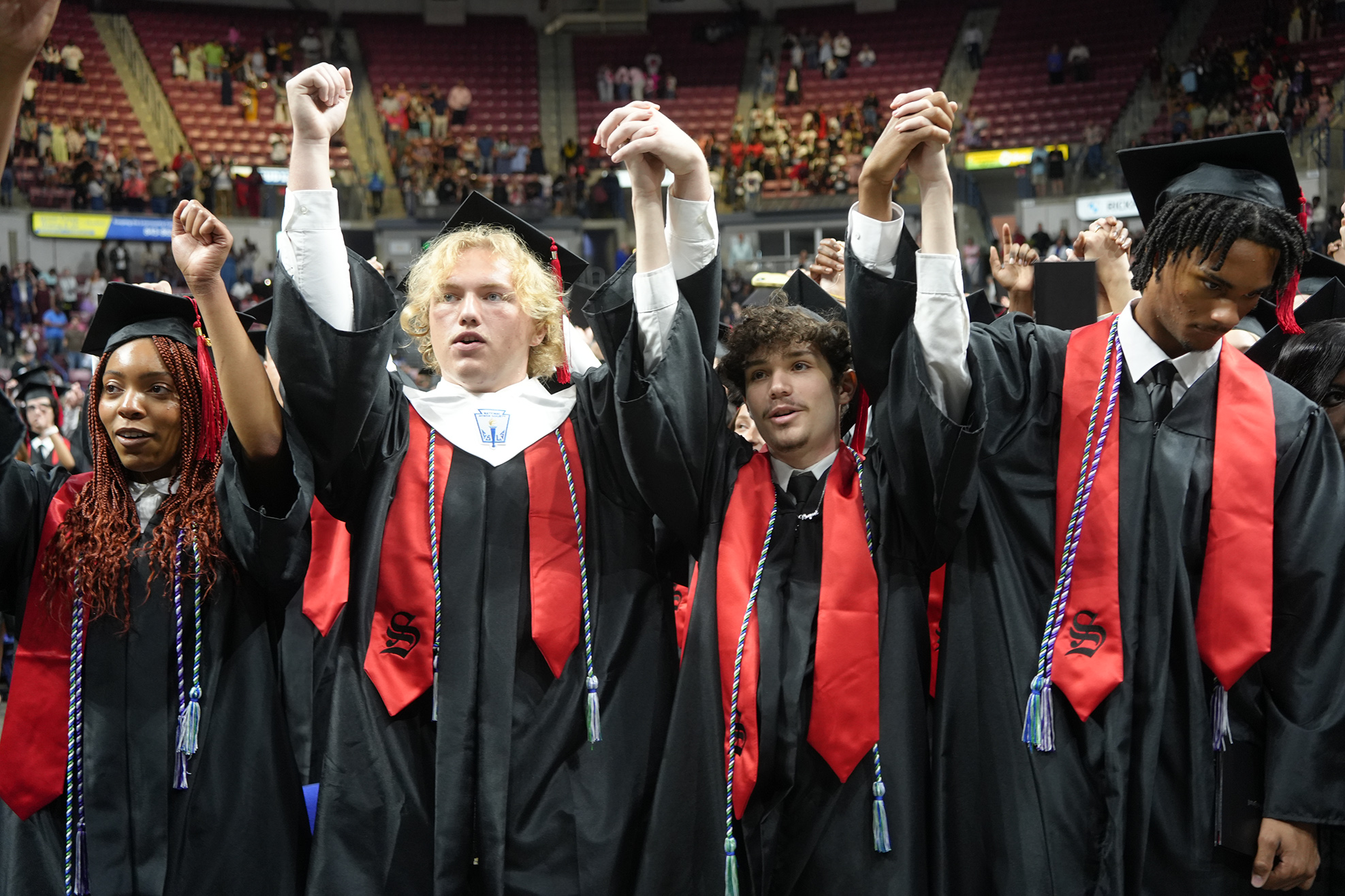 Group of individuals in black graduation gowns and caps raising their hands, standing in a row.