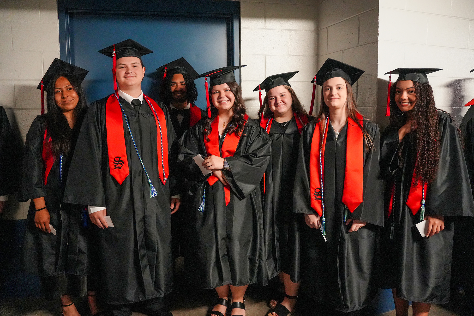 Graduates in caps and gowns stand together, smiling. They wear red stoles. A blue door is in the background.