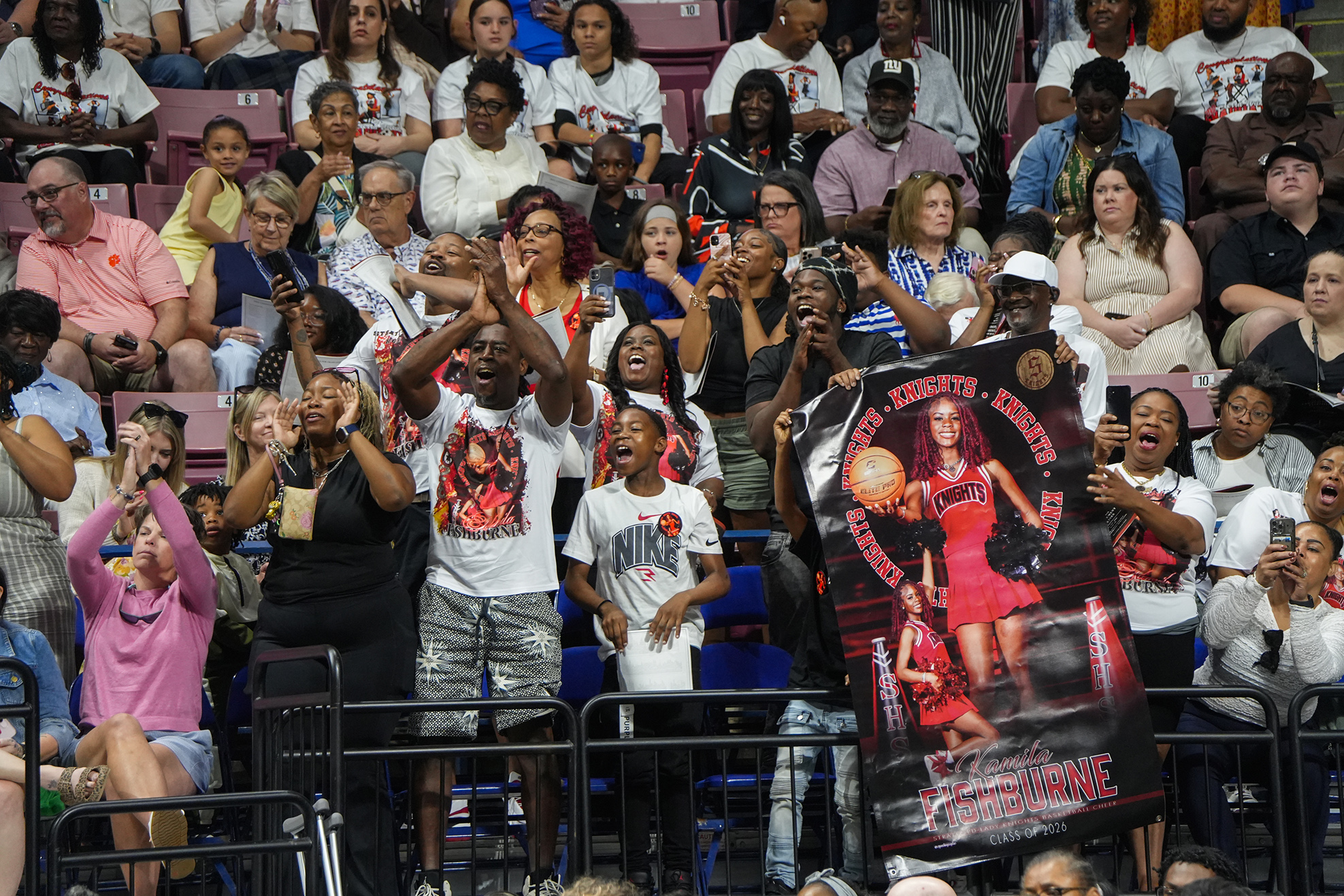 Spectators at an event, some cheering, with a banner featuring a sports figure in a red dress.