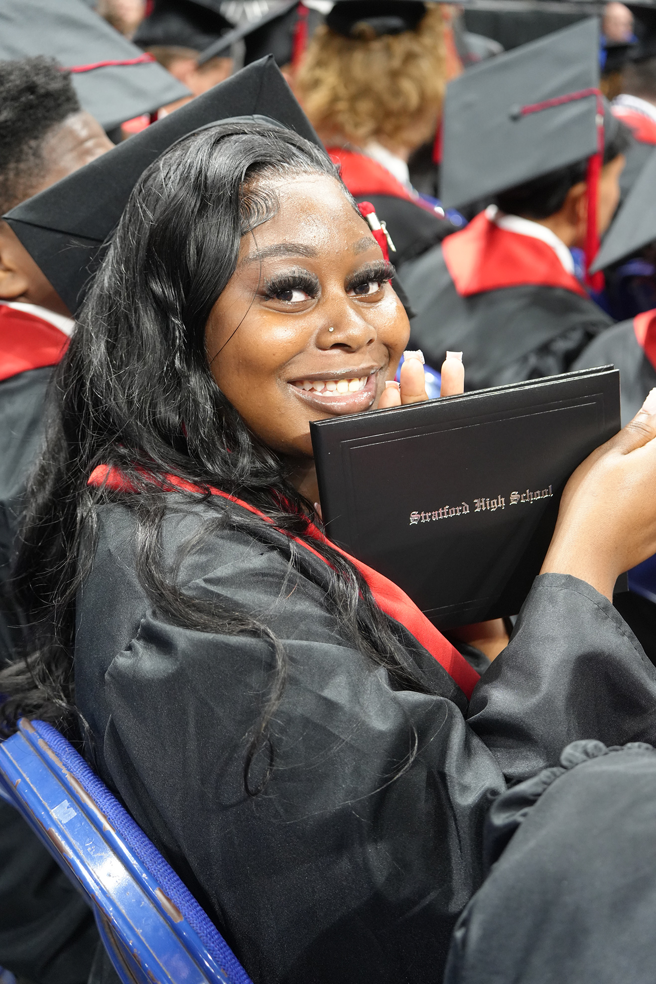 A woman wearing a graduation gown and cap smiles while holding a diploma. She sits among other graduates.
