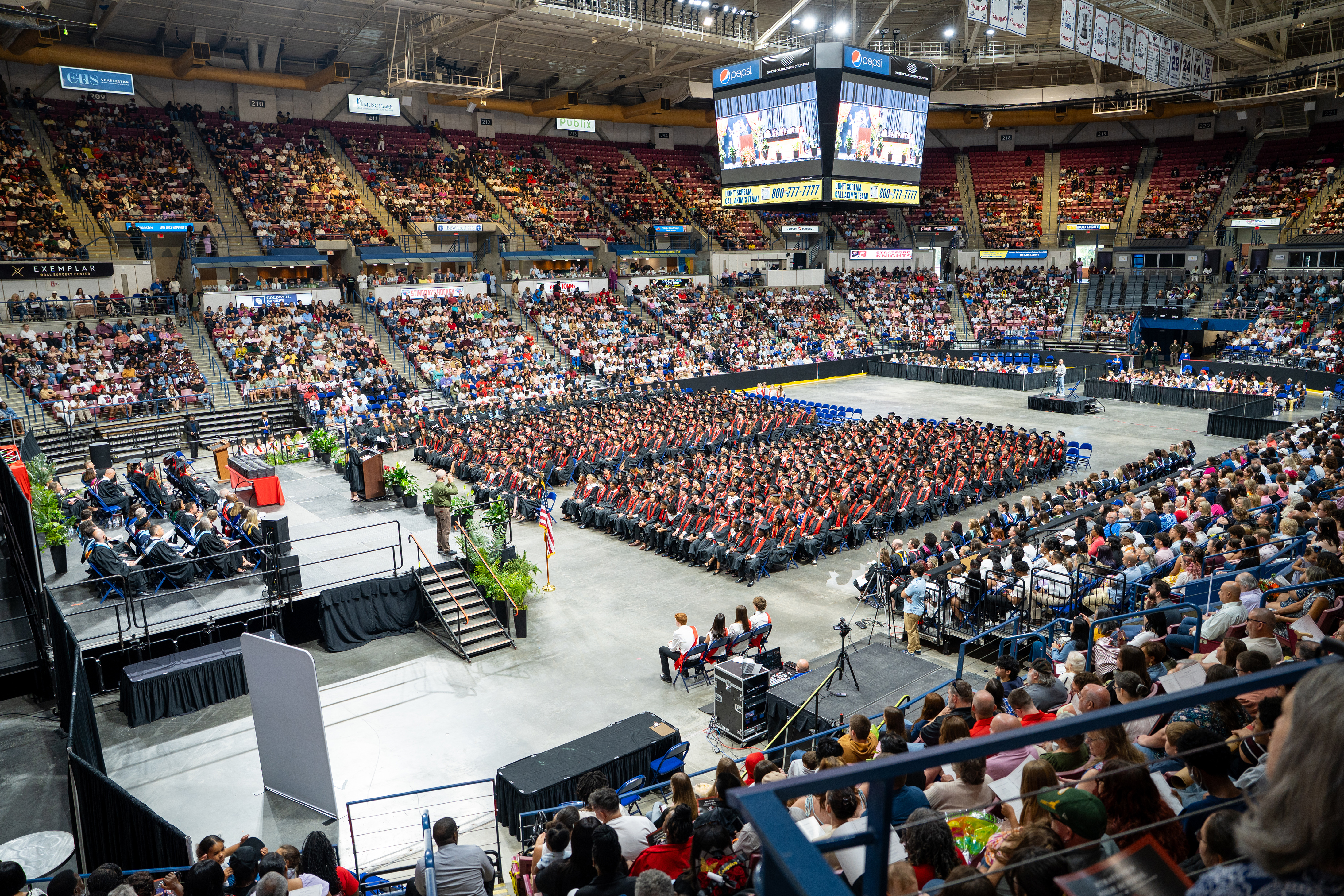 A large indoor stadium filled with people at an event, with seating areas, a stage, and a large screen.