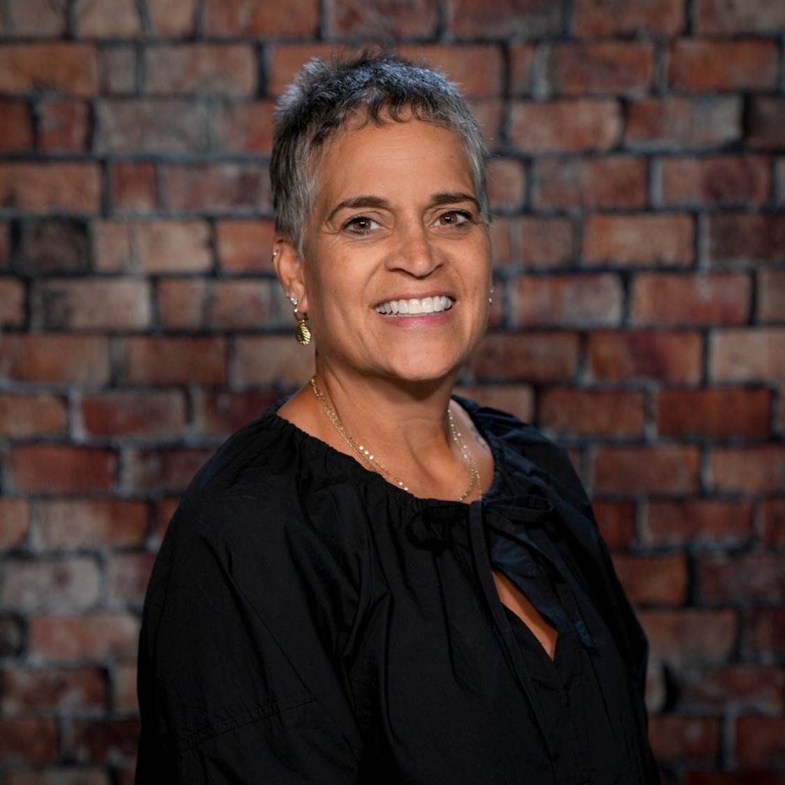 A smiling woman with short hair stands in front of a brick wall, wearing a black blouse and gold necklace.
