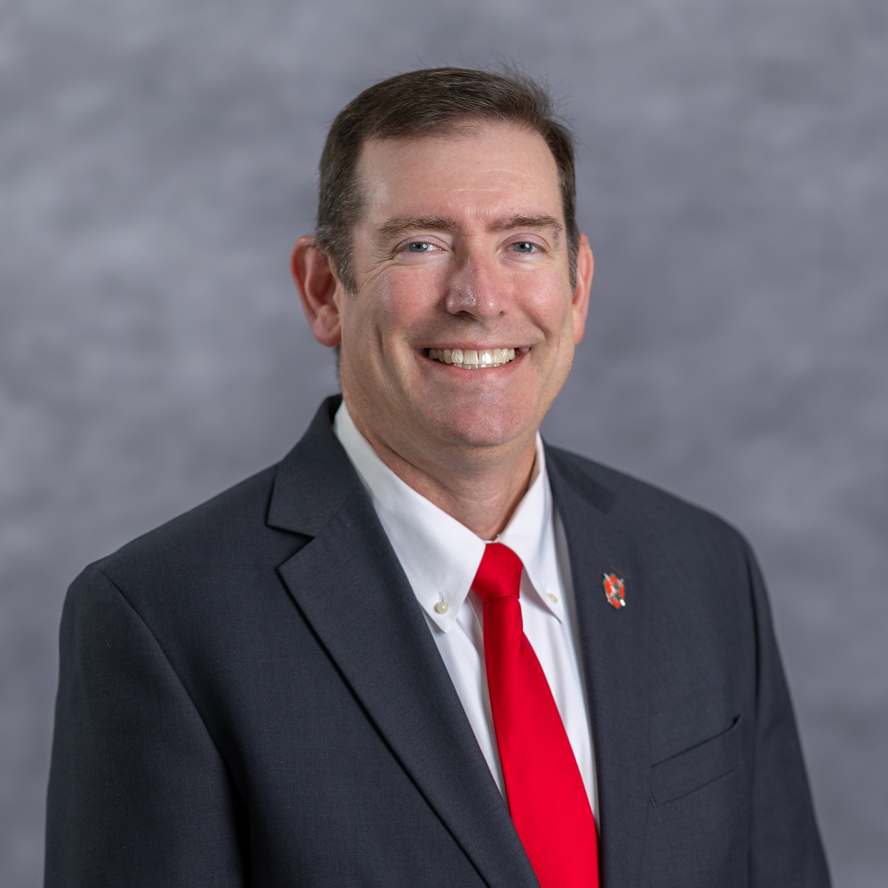 A man with short hair wearing a suit and red tie smiles, standing in front of a gray background.