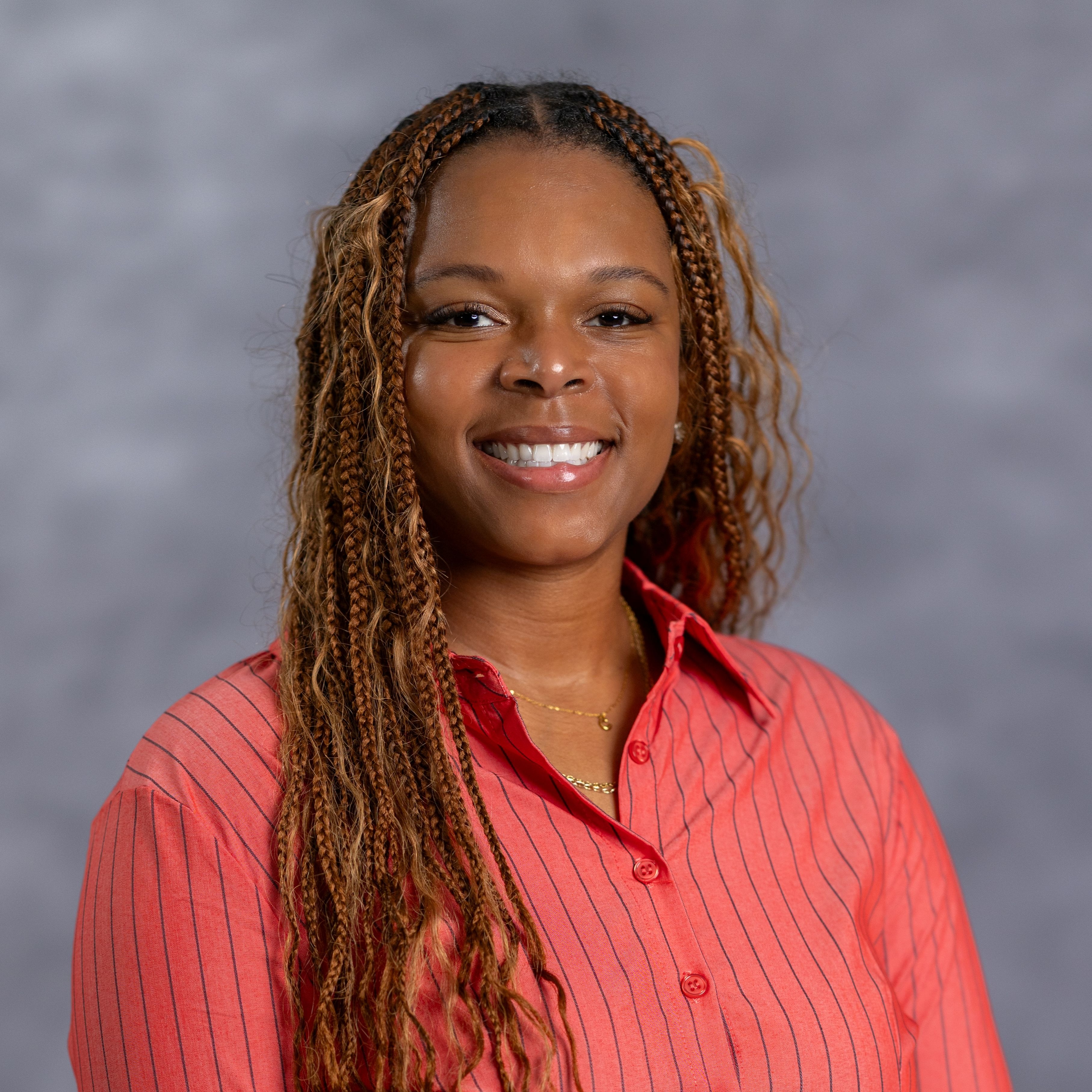 Woman with long, curly hair, wearing a red striped shirt, smiles at the camera. Background is plain and gray.