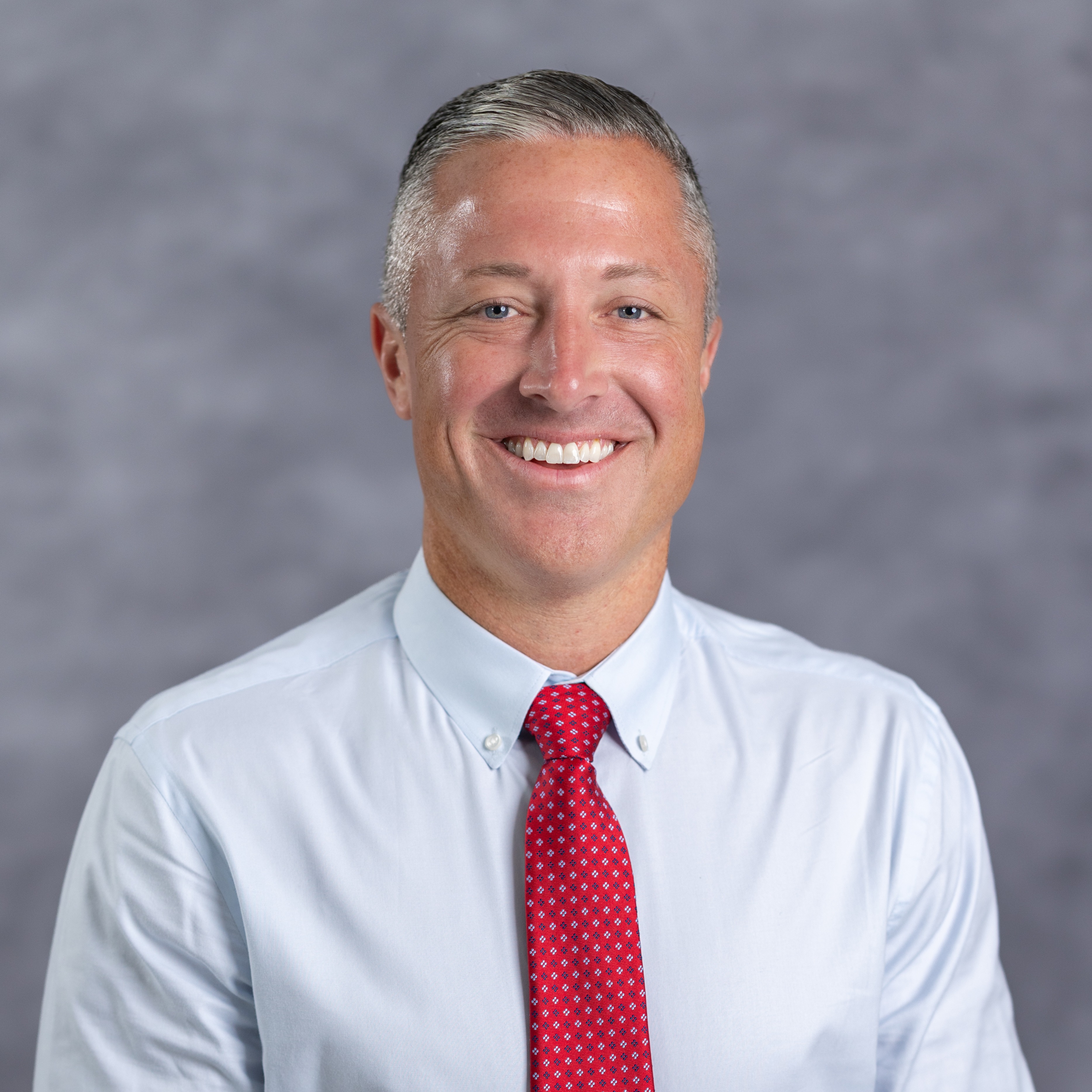 A man stands smiling with a light blue shirt and a red tie with white polka dots. The background is solid gray.