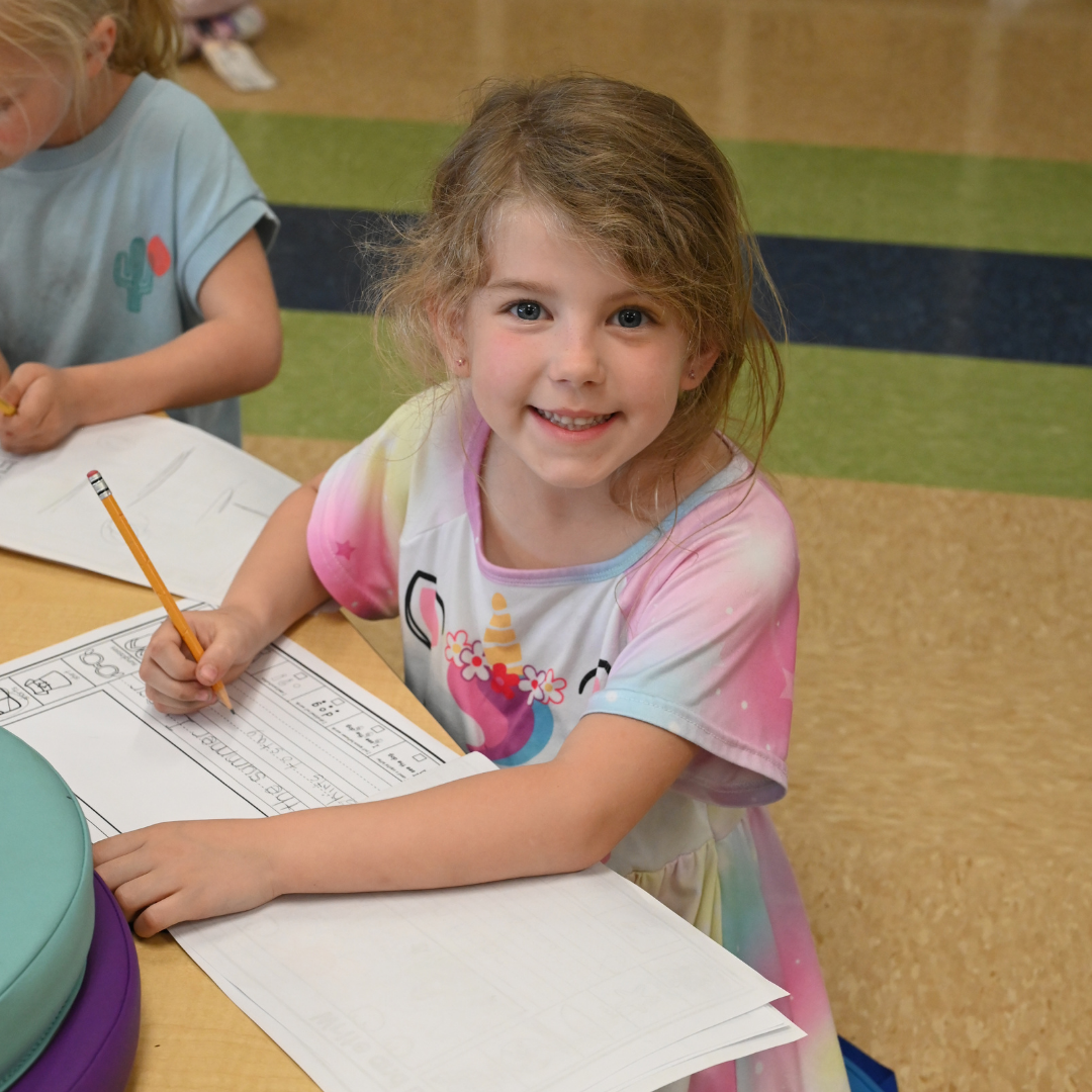 Student writing on paper with a pencil while smiling at the camera.