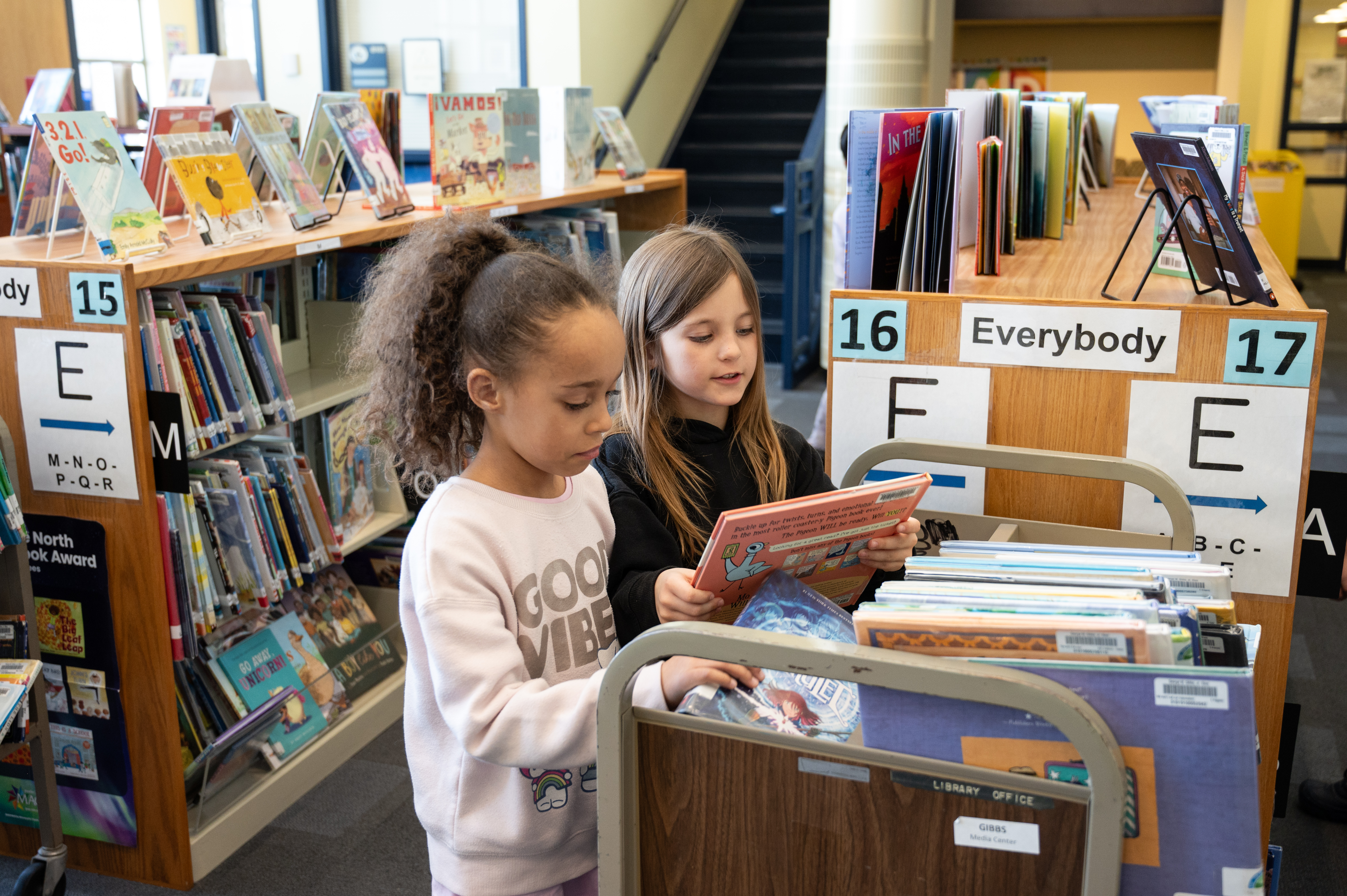 Two girls browse a library cart looking for their next favorite read.