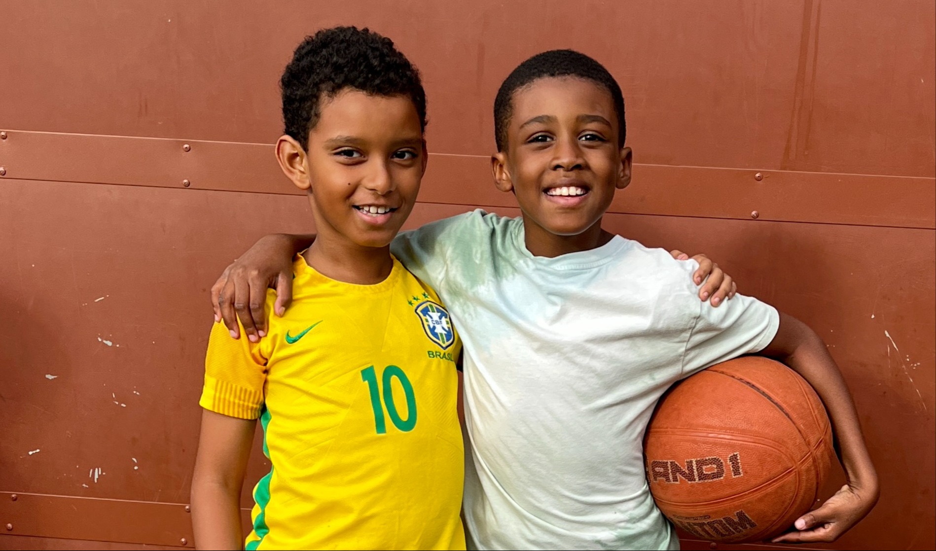 Two boys smiling, arm in arm, one holding a basketball