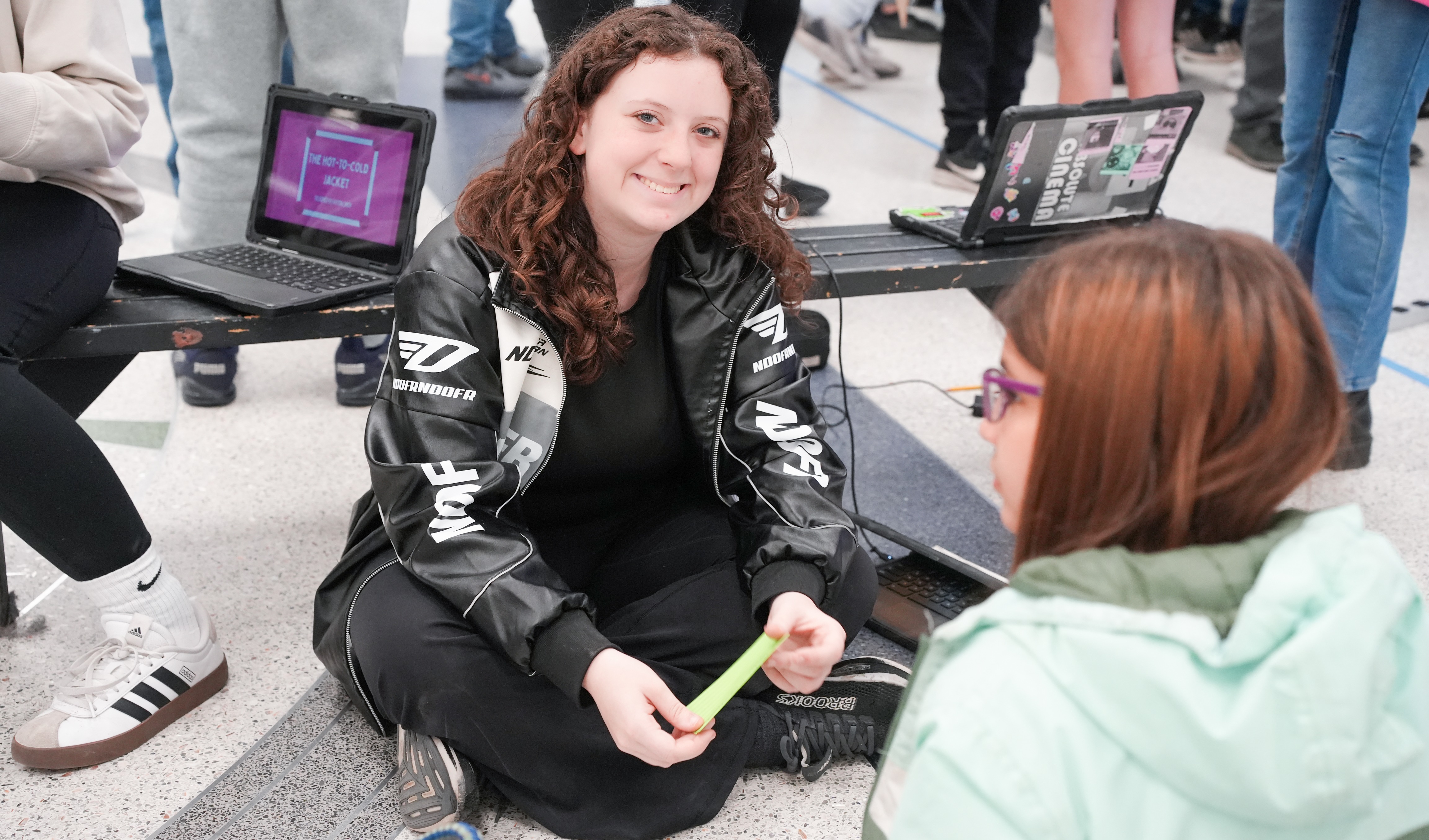 A woman in a black jacket sits on a floor, holding a green object. She converses with a seated woman wearing a green jacket.