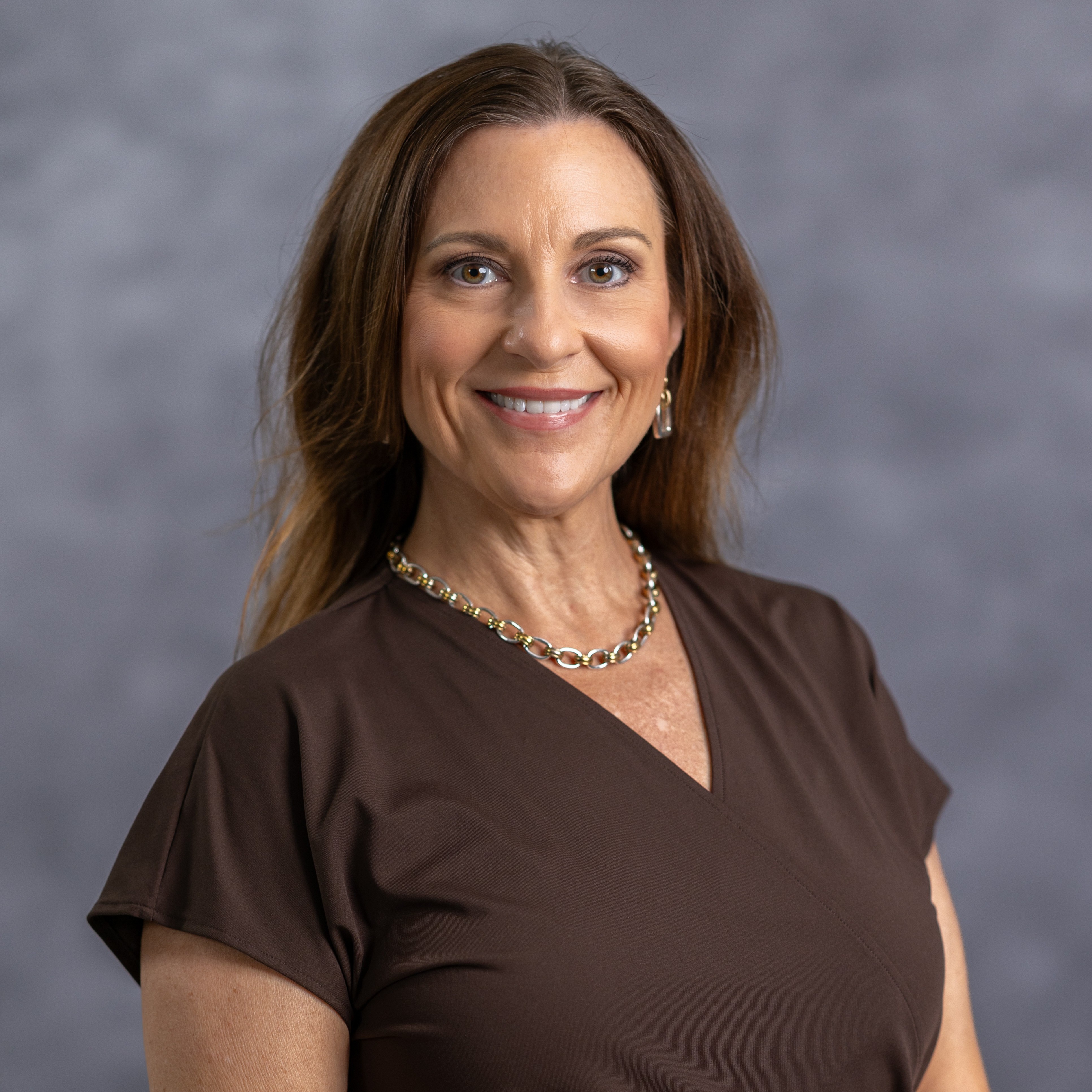 A smiling woman wearing a brown top and necklace. She has long hair and is standing against a gray background.