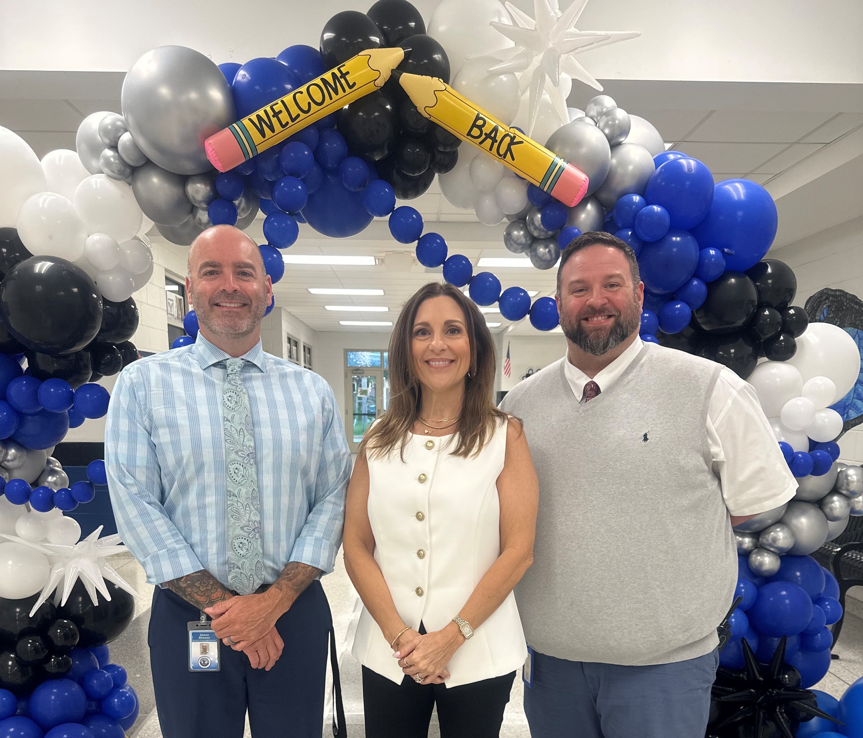 Three people pose in front of a colorful balloon arch with a "Welcome Back" theme.