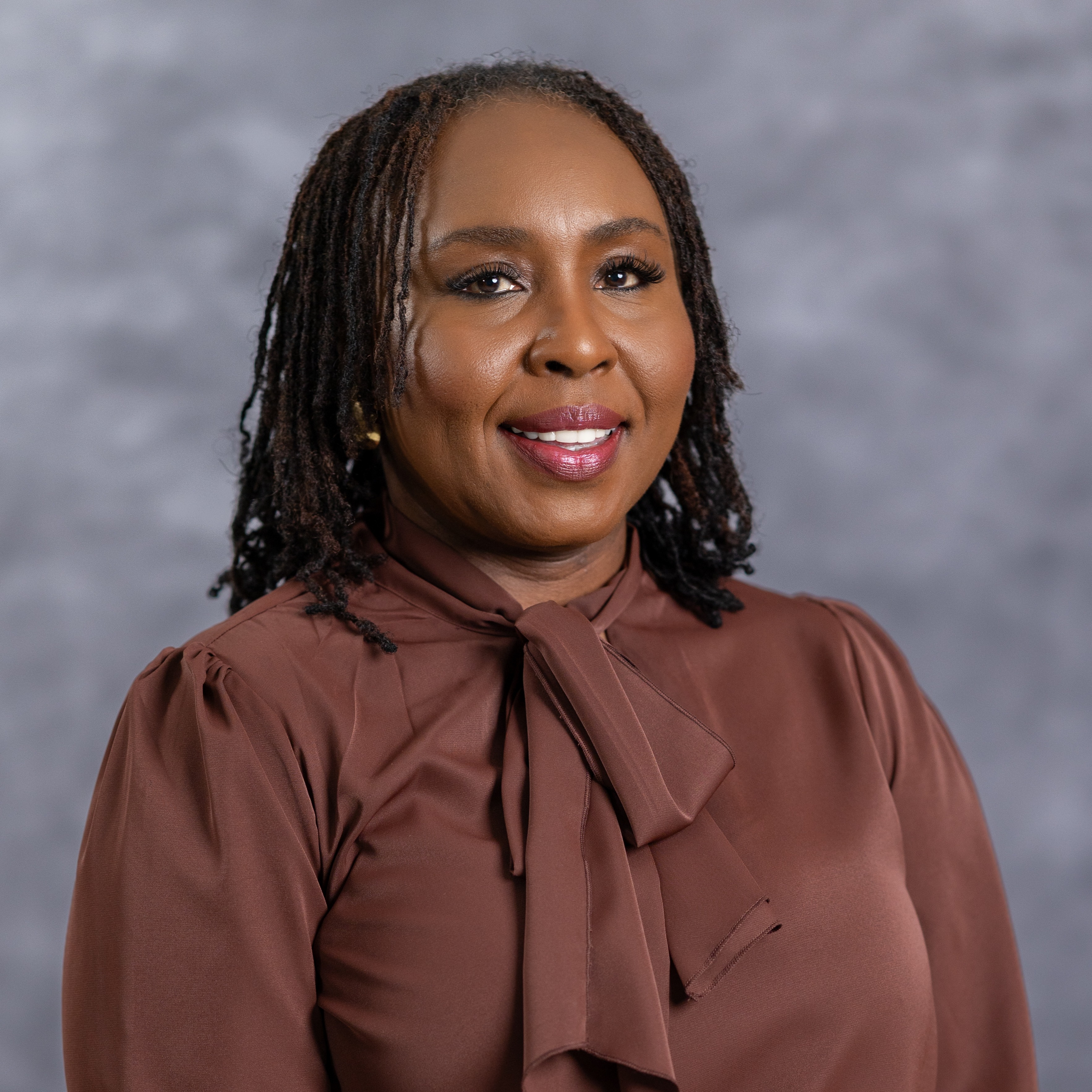A woman with long, braided hair wearing a brown blouse with a bow at the neck.
