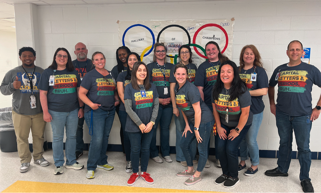 english teachers stand before a cinderblock wall with a mural of the olympic rings. teachers wear matching shirts proclaiming, "capital letters & periods, bruh"
