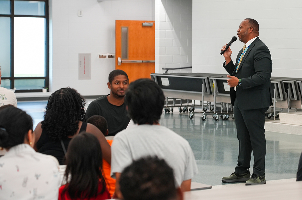 A man speaks into a microphone while standing in a room with chairs, and others sit in chairs, listening.