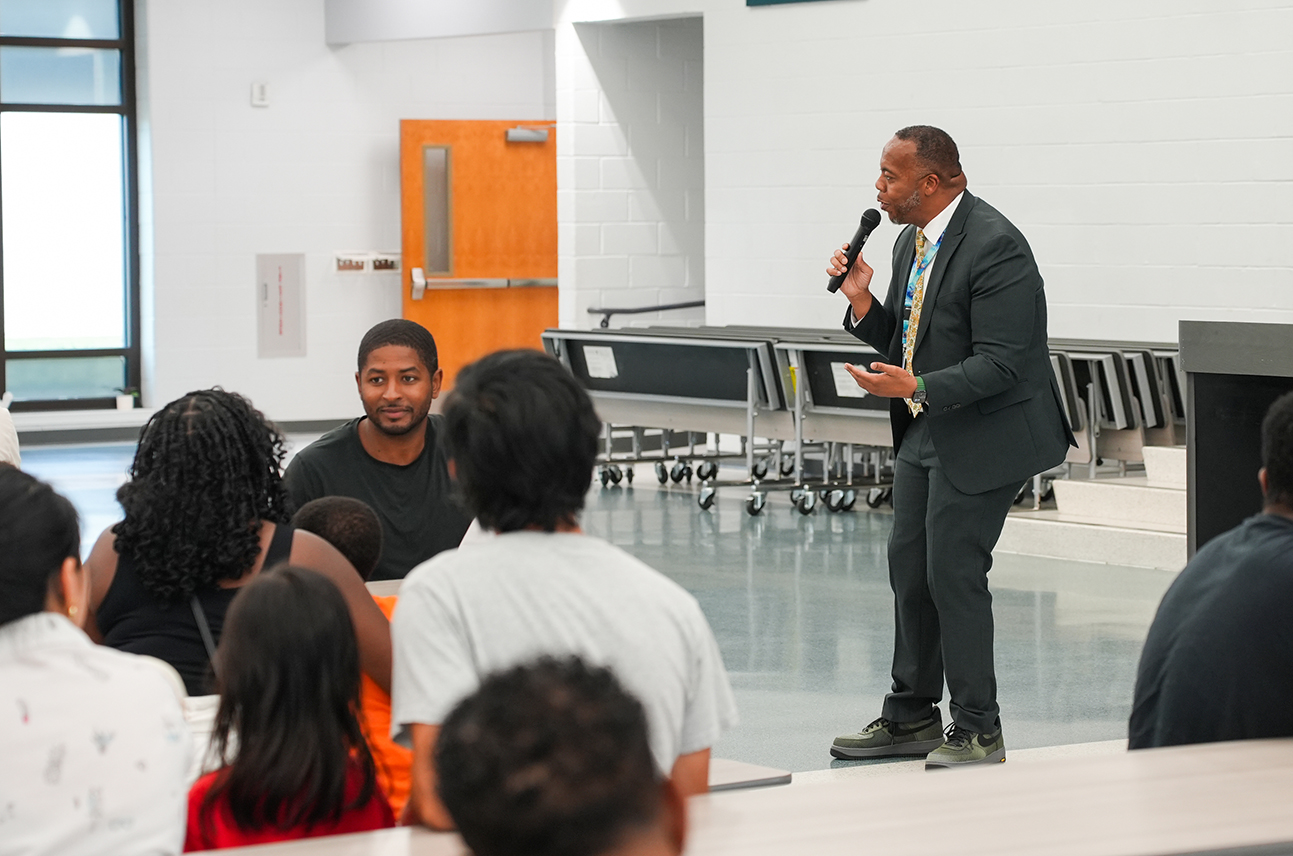 A man stands before a group in a classroom, speaking into a microphone, with attendees seated.
