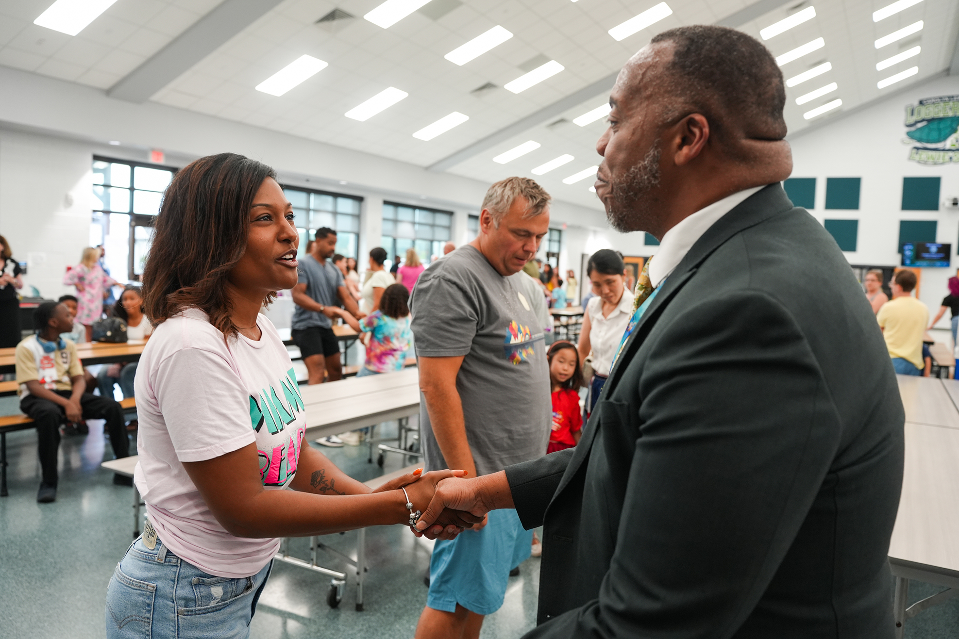A woman and man shake hands in a room with others; they wear casual and formal attire.