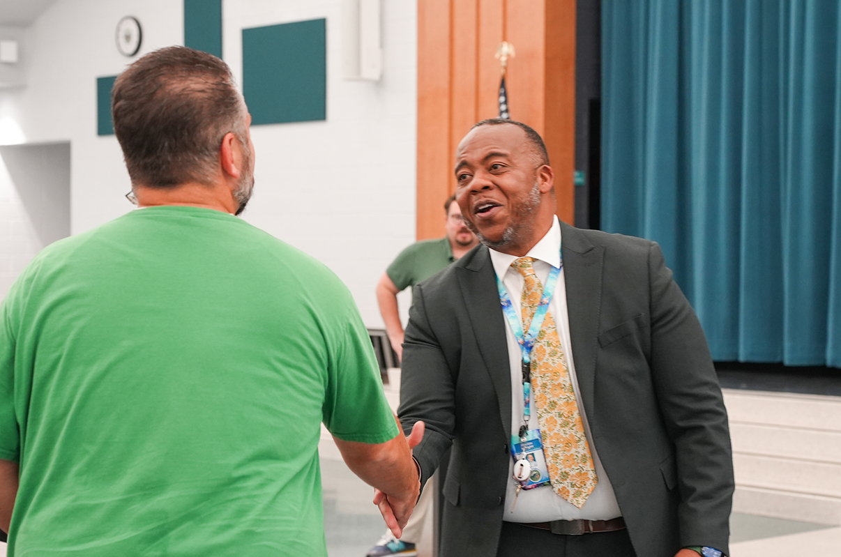 Two men shake hands in an indoor area. One wears a green shirt and the other a suit with a yellow tie.