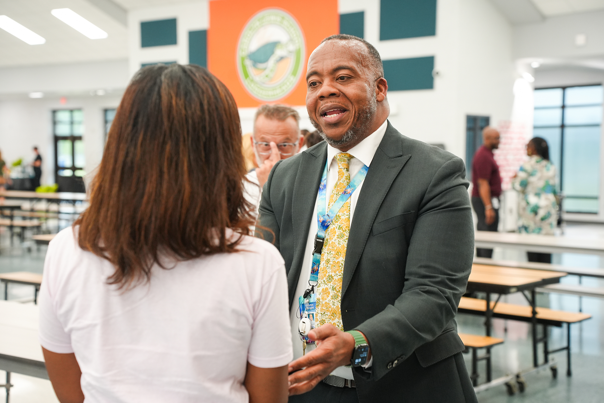 A man in a suit speaks to a woman in a cafeteria. Behind them, two people stand. Bright lights illuminate the area.