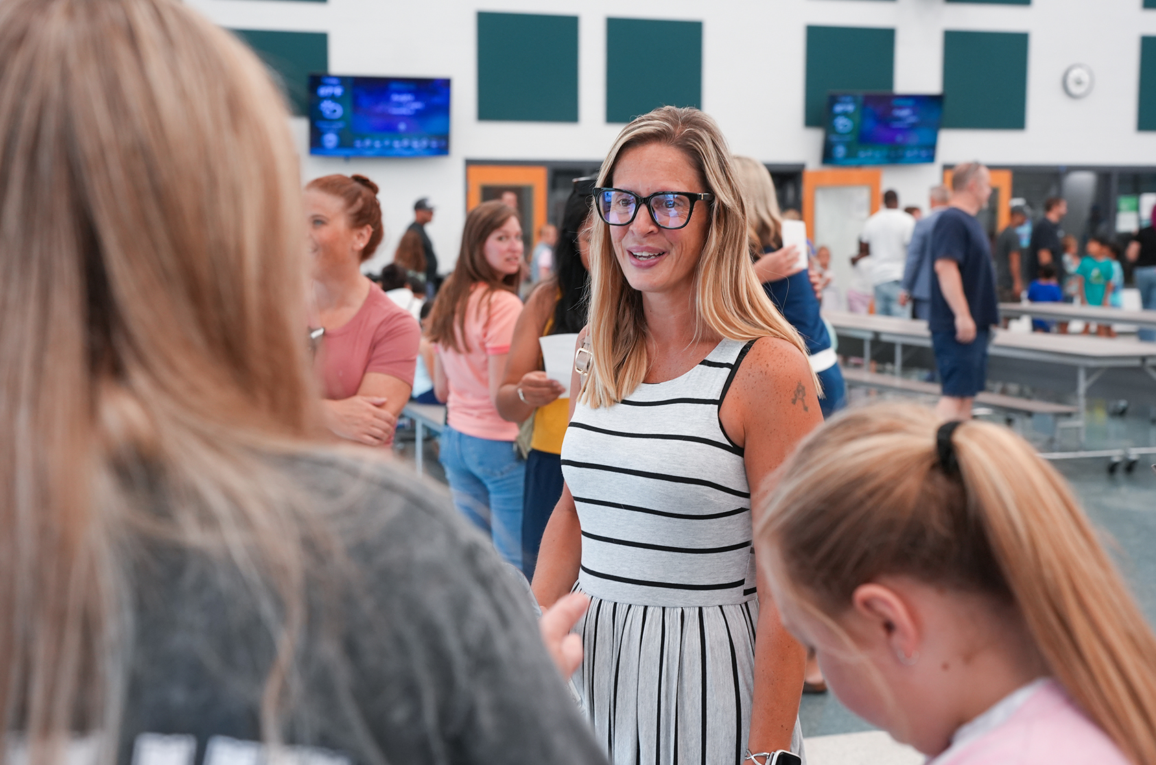 A woman in glasses and a striped dress speaks to a girl and another woman in an indoor area.
