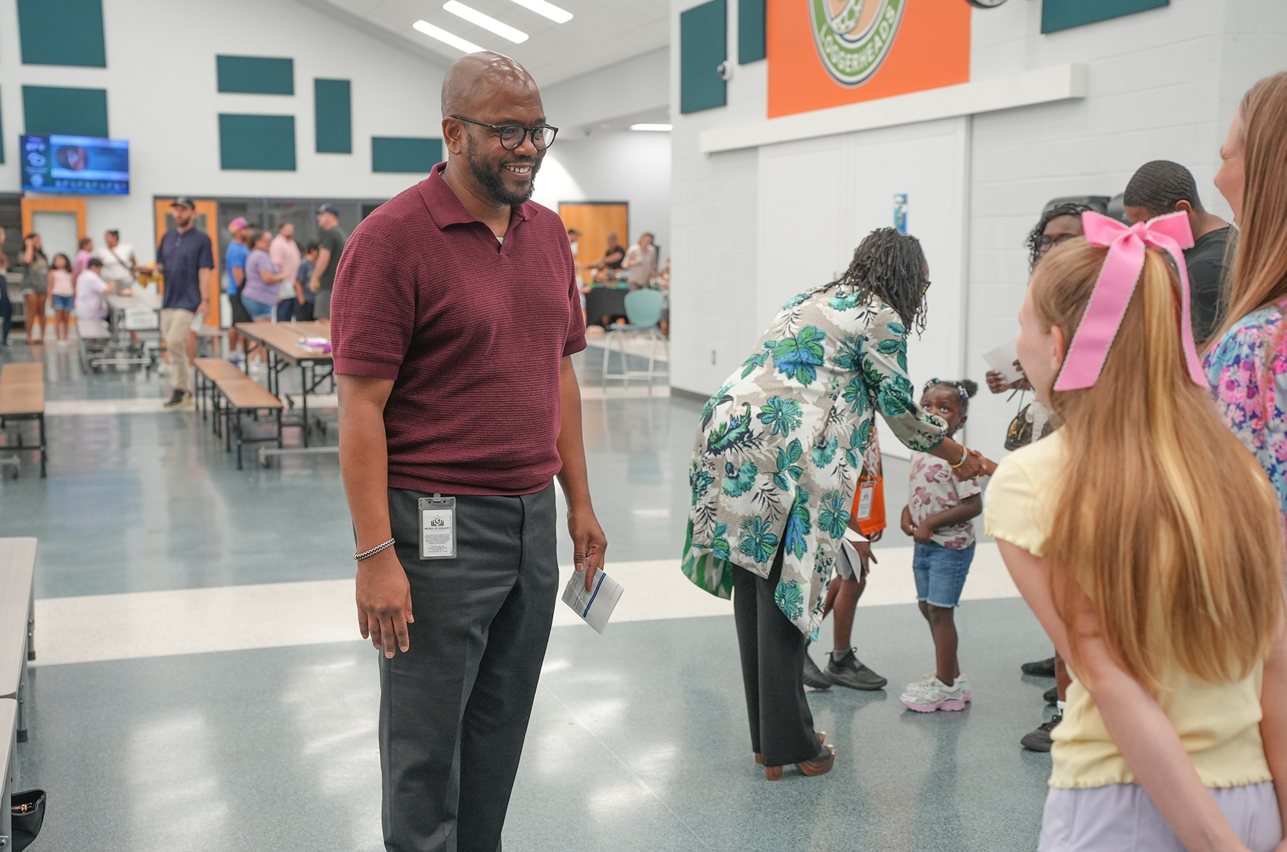 Adults and children in a cafeteria with white walls and orange accents. One man talks to a young girl.