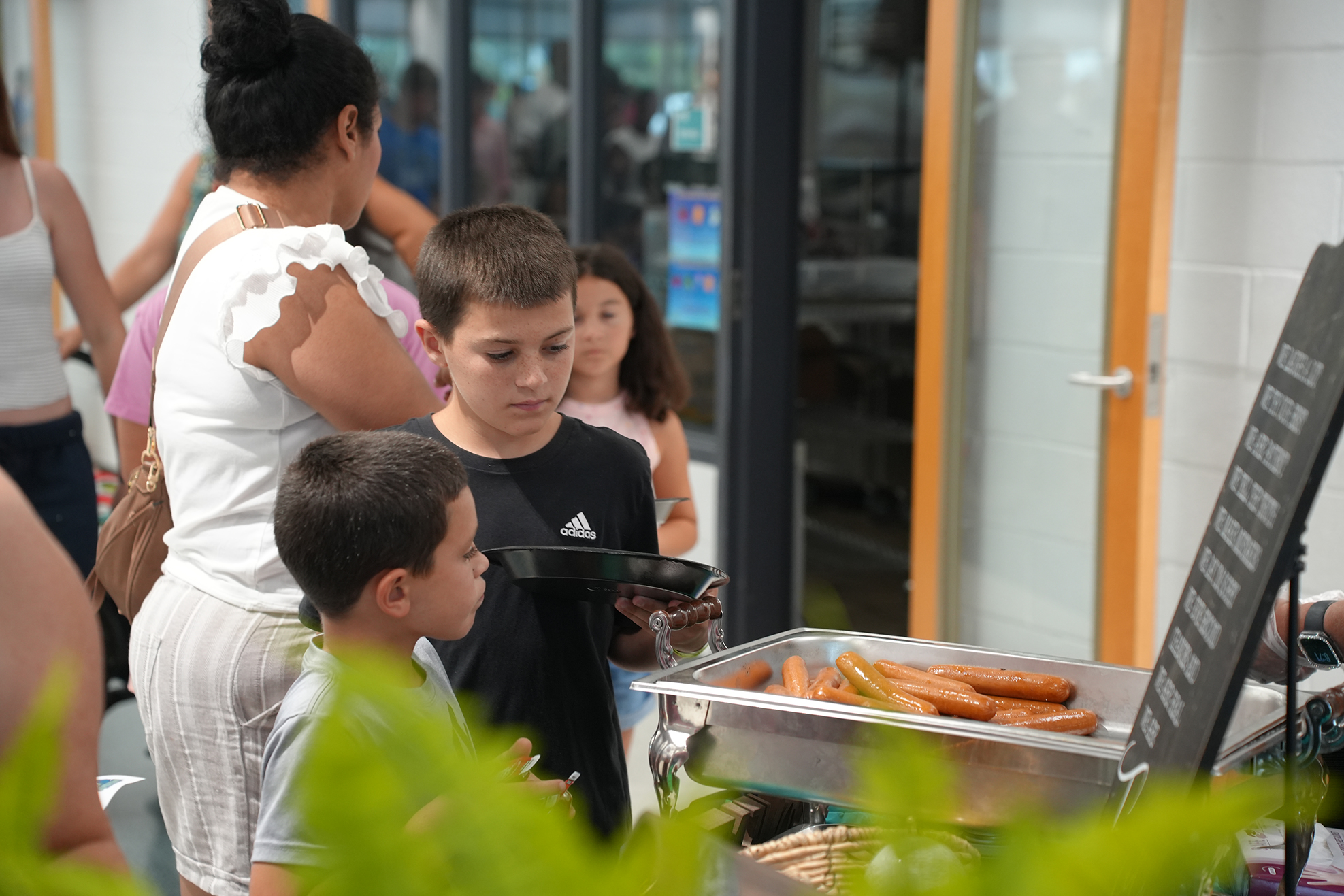 A group of people stand at a food station with hot dogs in trays, while a woman holds a tray.