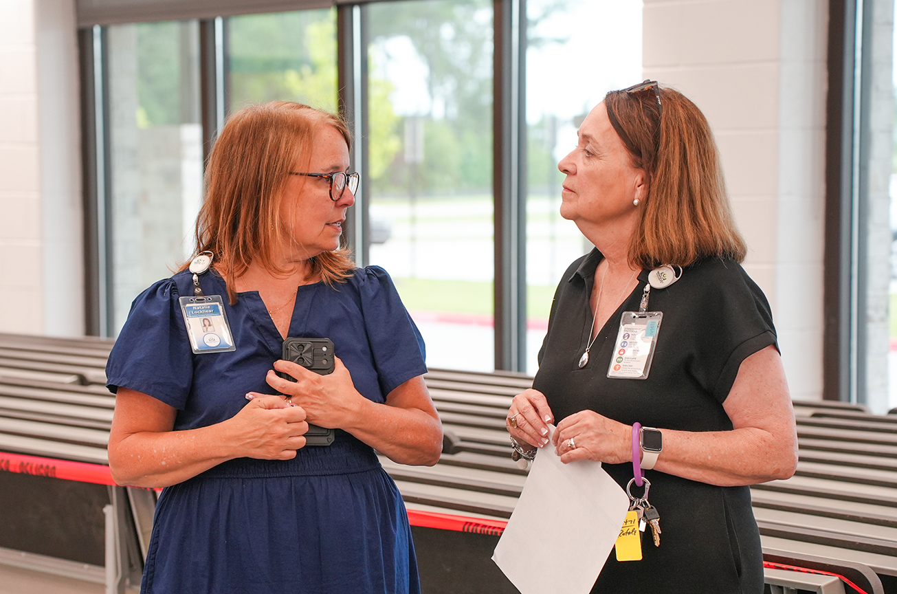 Two women speak, one in blue with a badge, the other in black with a paper. Behind them, a large glass window shows outdoor trees.