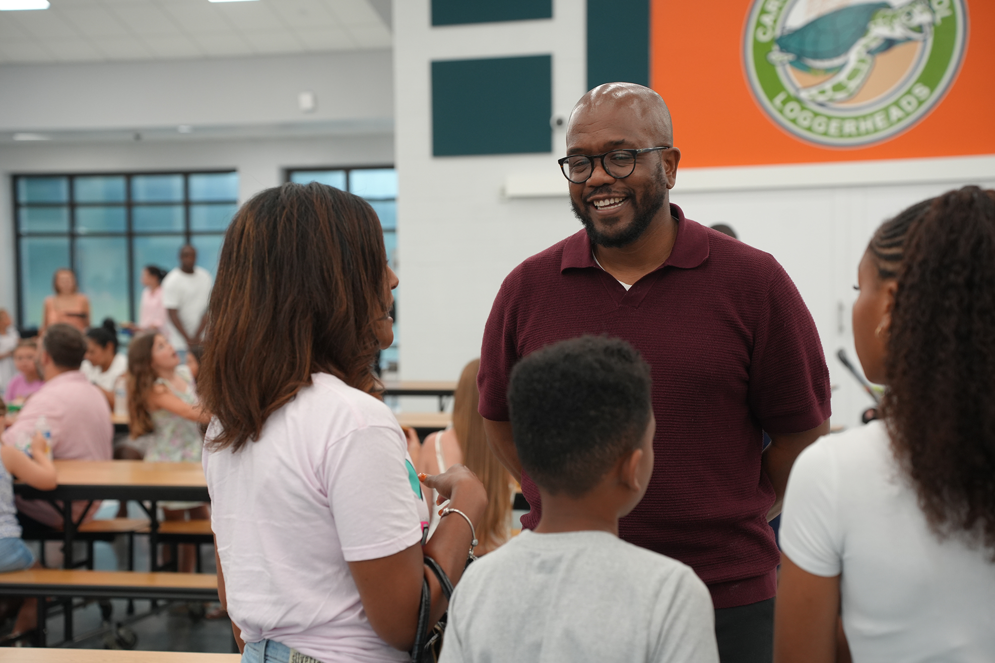 A man and three women stand facing each other in a cafeteria. The man smiles and wears glasses.