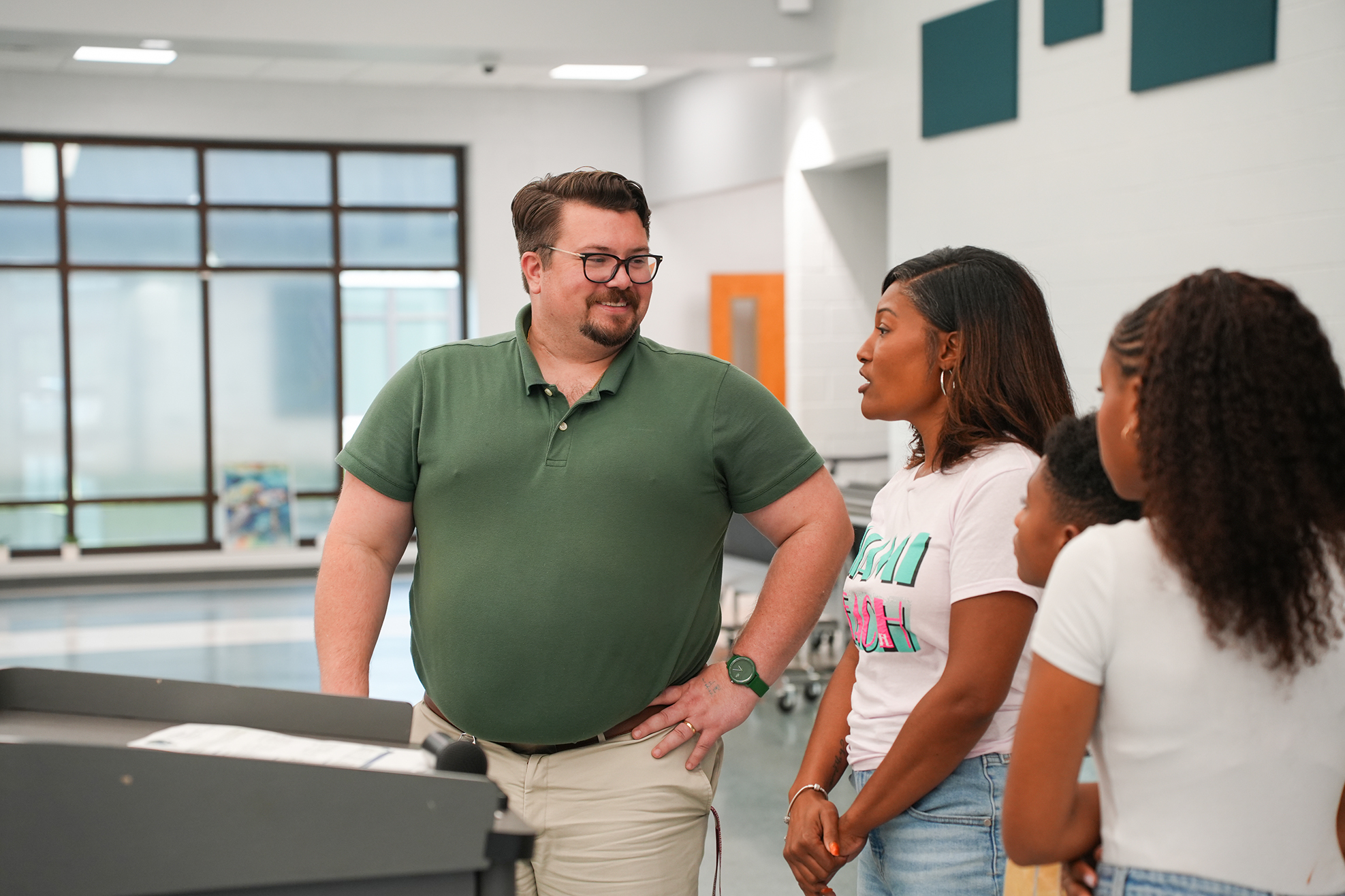 A man in a green polo shirt and glasses speaks to two women and a girl in a room with windows.