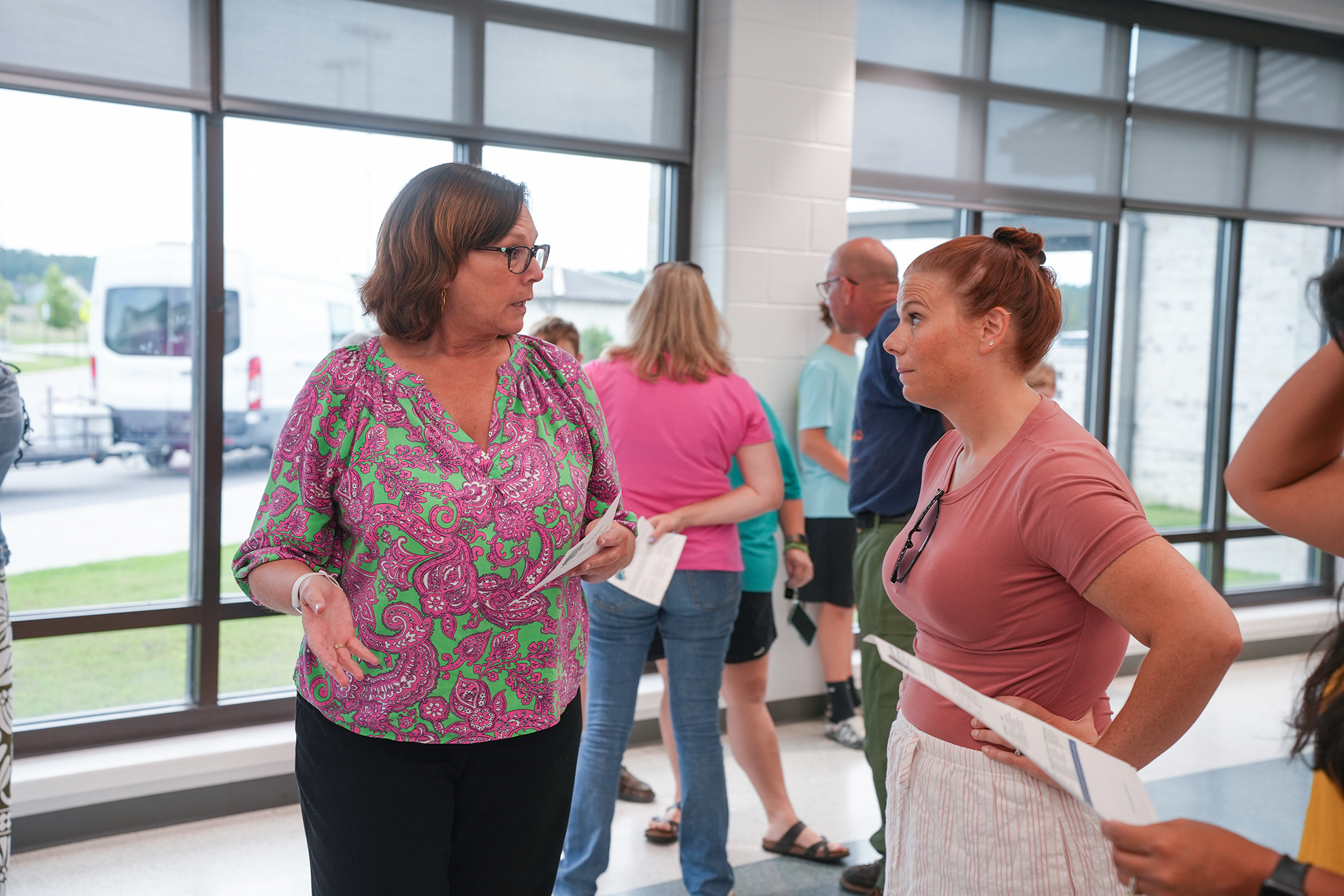 Several people inside a room with glass walls. Two women face each other, holding papers. Others stand nearby.