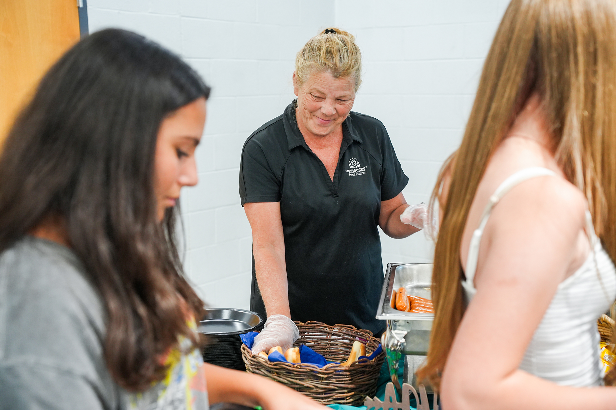 Three women in a room. One serves food from a basket, another holds a plate. The third watches.