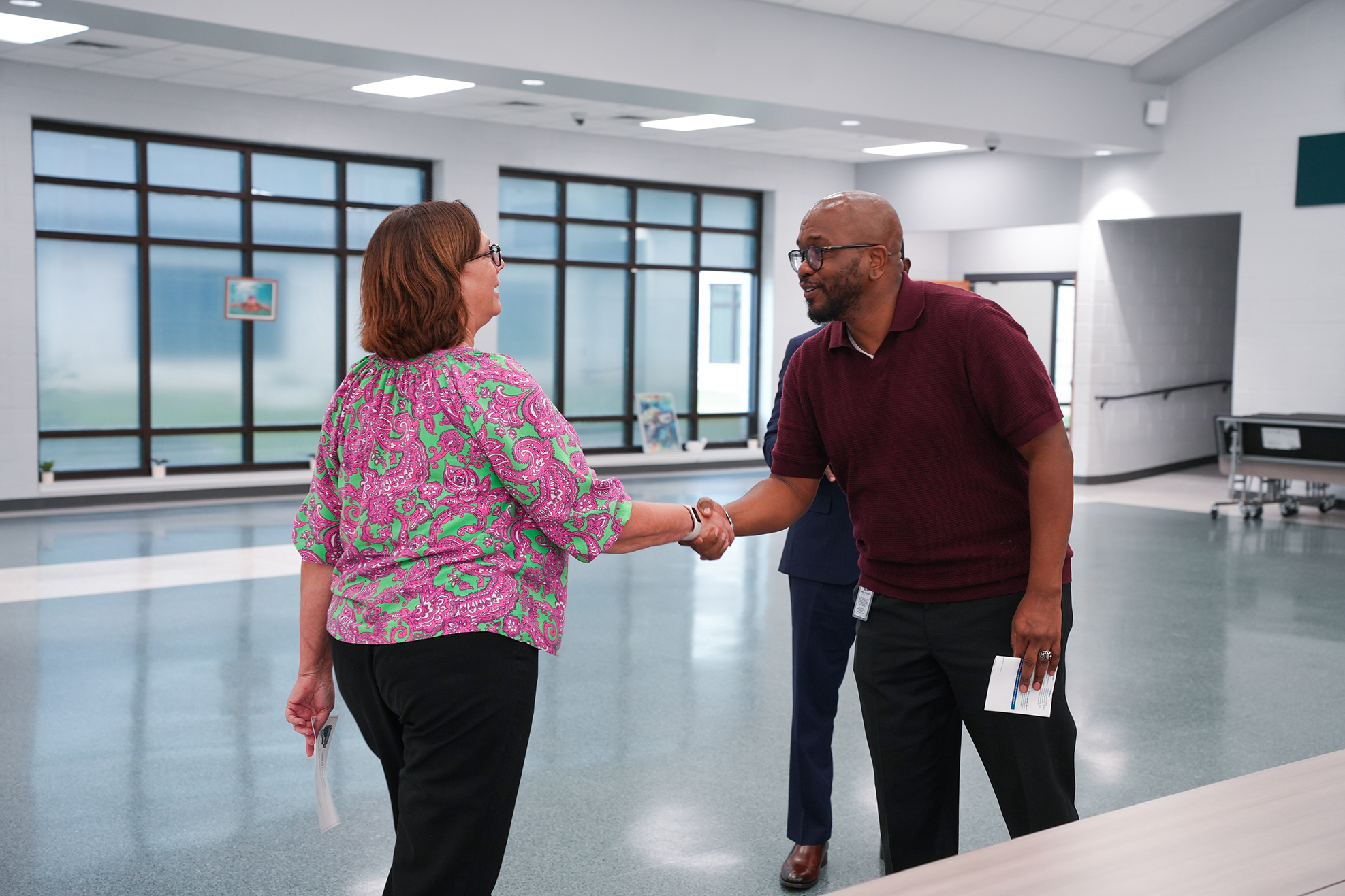 Two people in a large room with bright lights shake hands. One holds a folder. Behind them, another person stands.