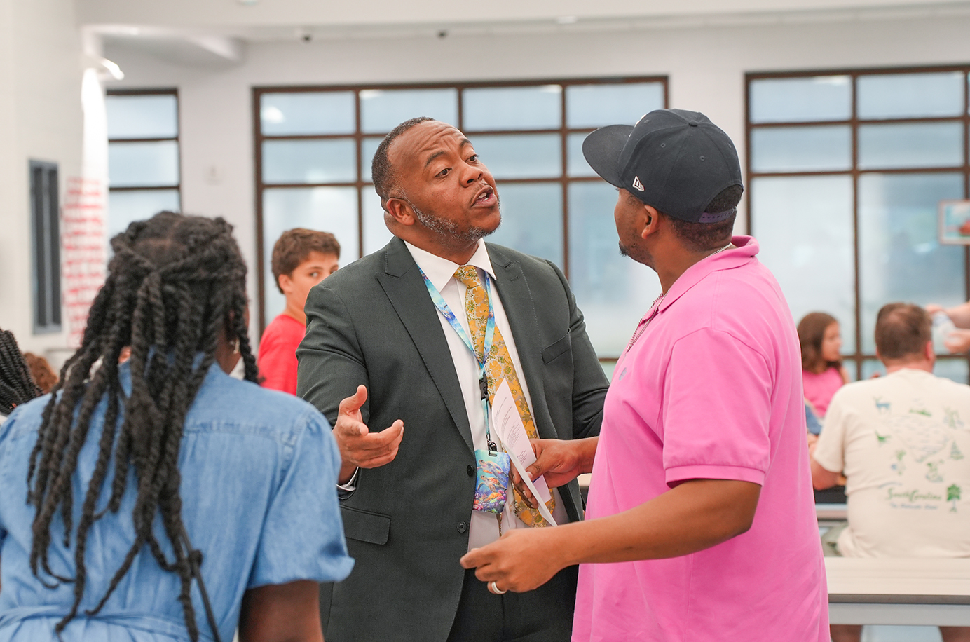 A man in a suit speaks to two others in a classroom. The woman listens.