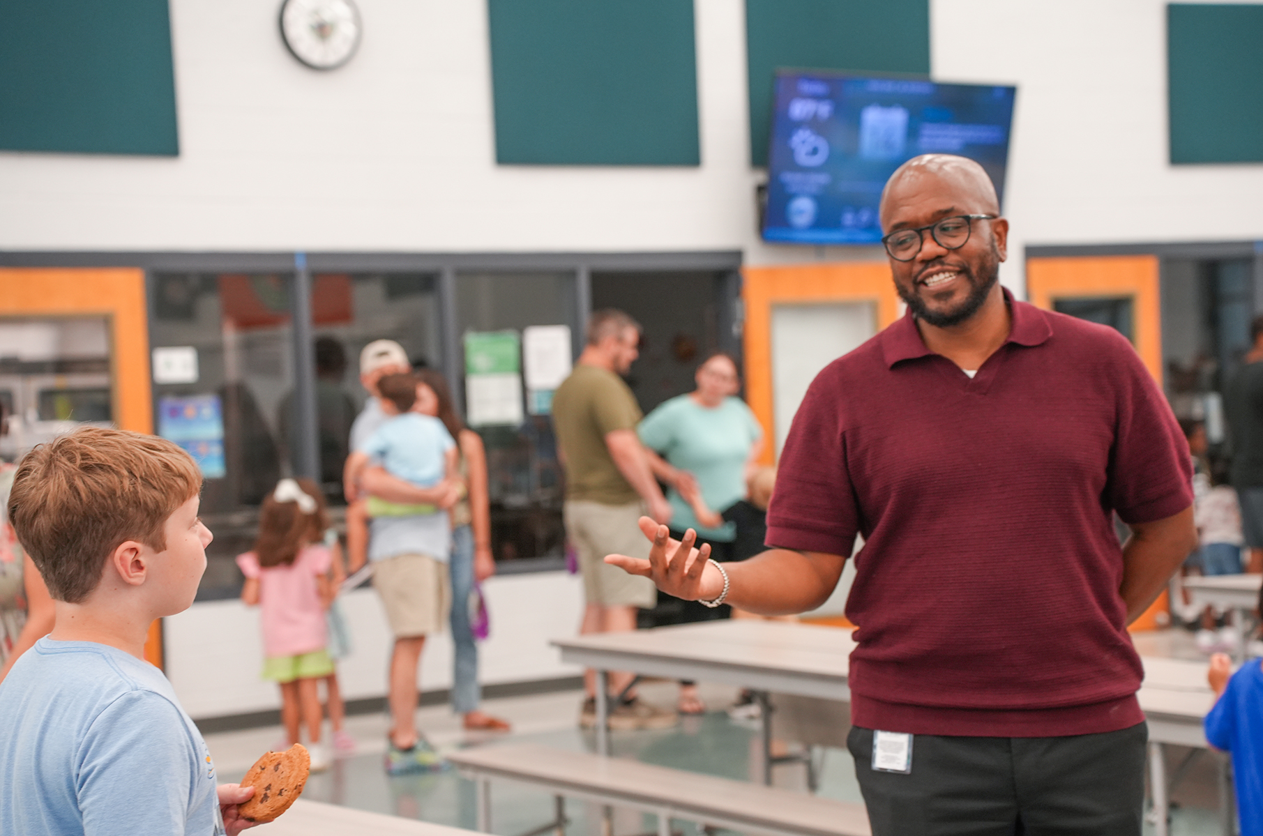 A man in a maroon polo speaks to a child while holding a cookie. They are in a dining area with tables.