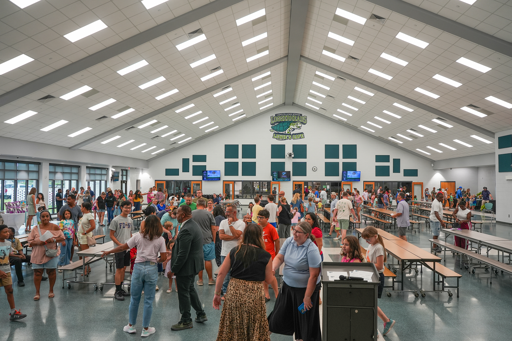Indoor cafeteria with large tables and benches, ceiling lights, and a diverse group of people.
