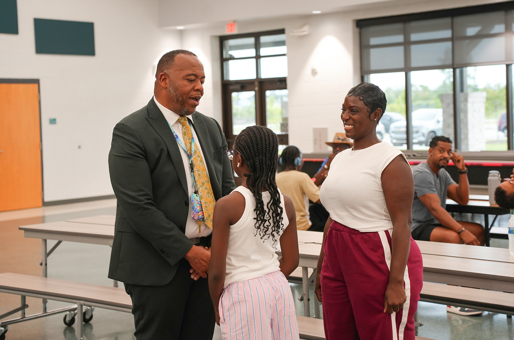 A man in a suit speaks with a woman and a girl in a cafeteria with white walls.