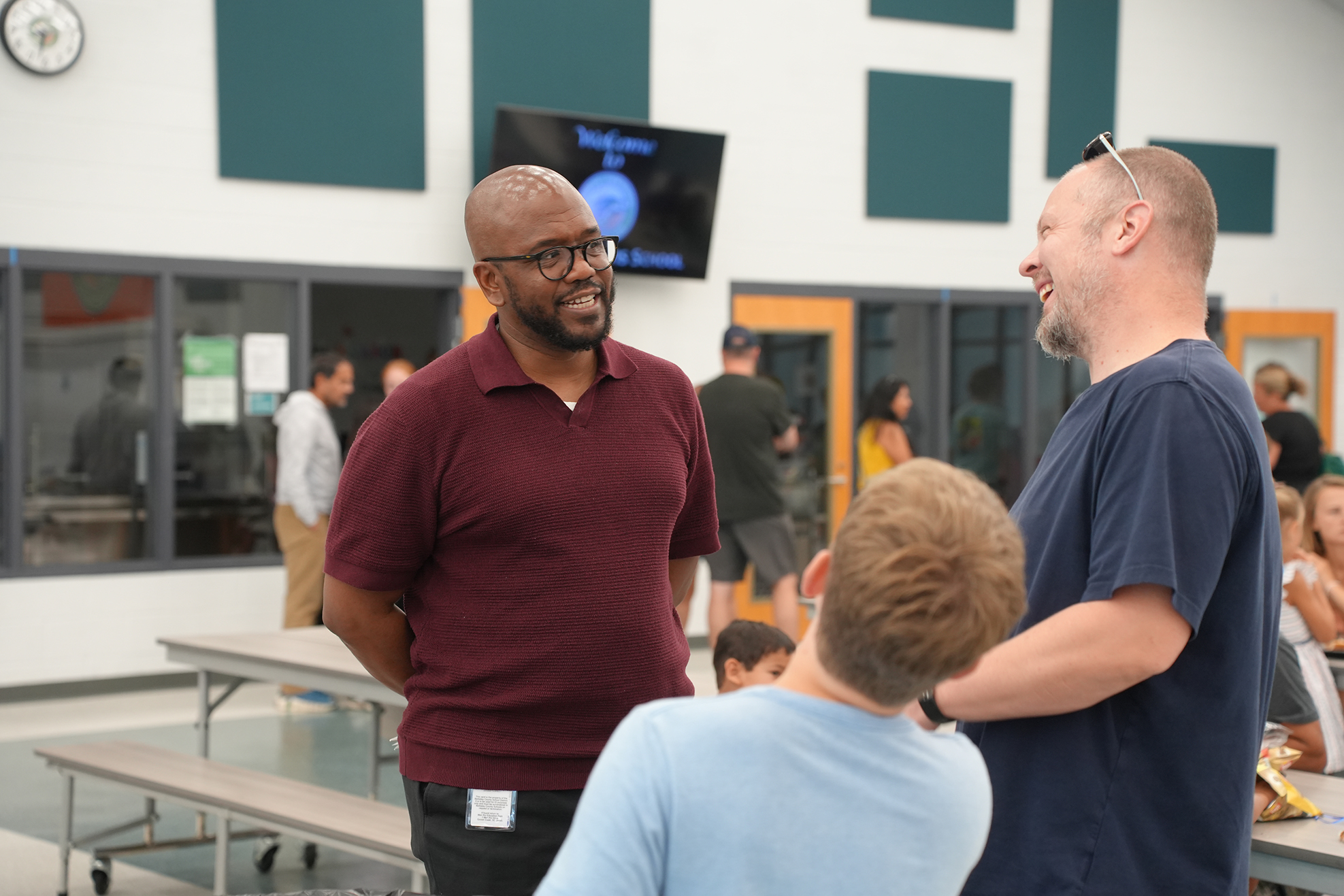 Two men and a boy converse in a school cafeteria with tables, a monitor, and other people.