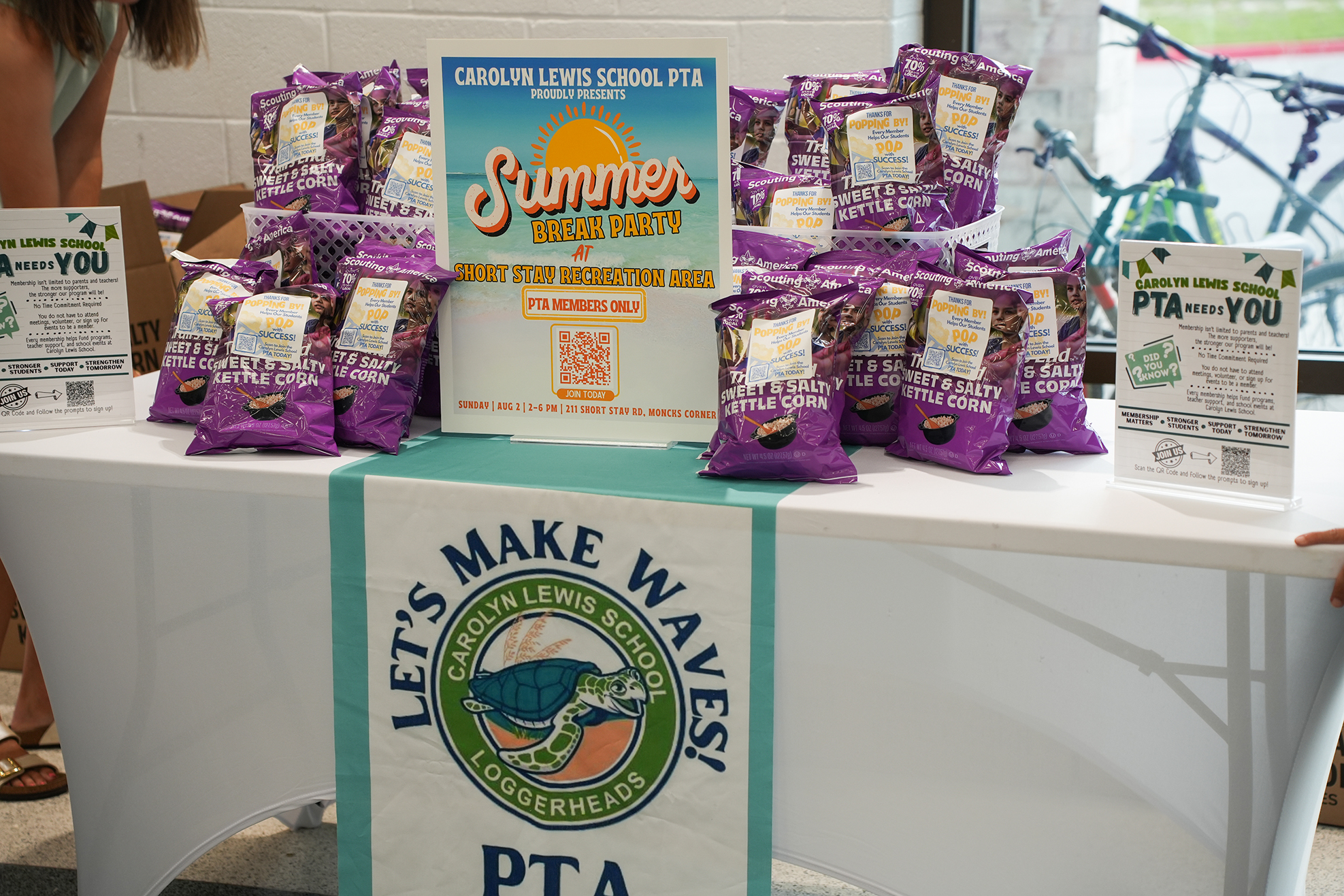 A white table displays bags of purple snacks. A blue and white banner reads "Let's Make Waves" and "PTA".