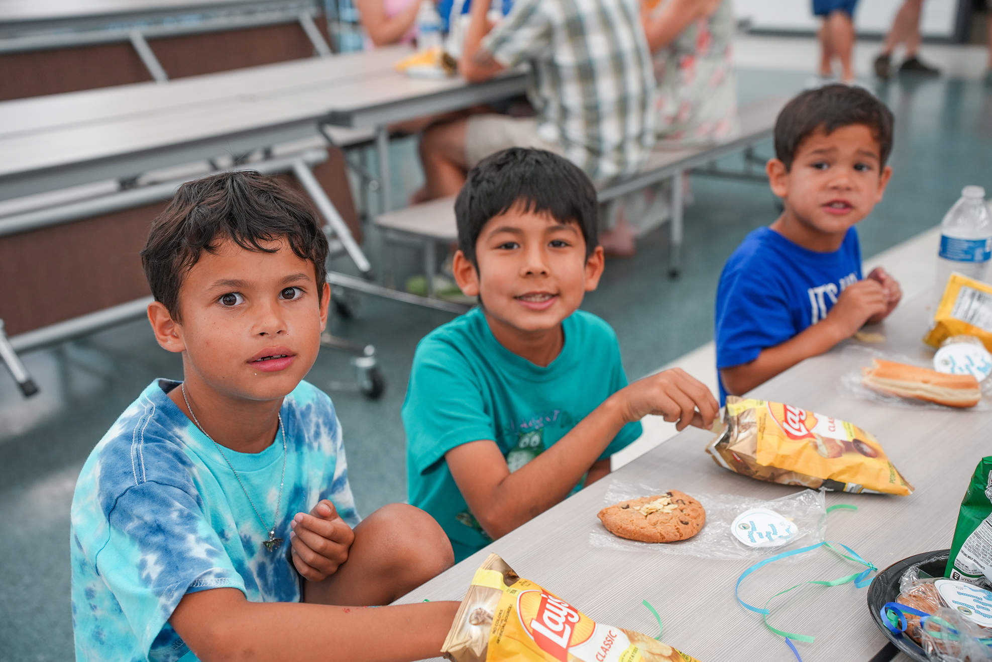 Three boys in a cafeteria. One eats cookies, another eats chips, and a third wears a blue shirt.