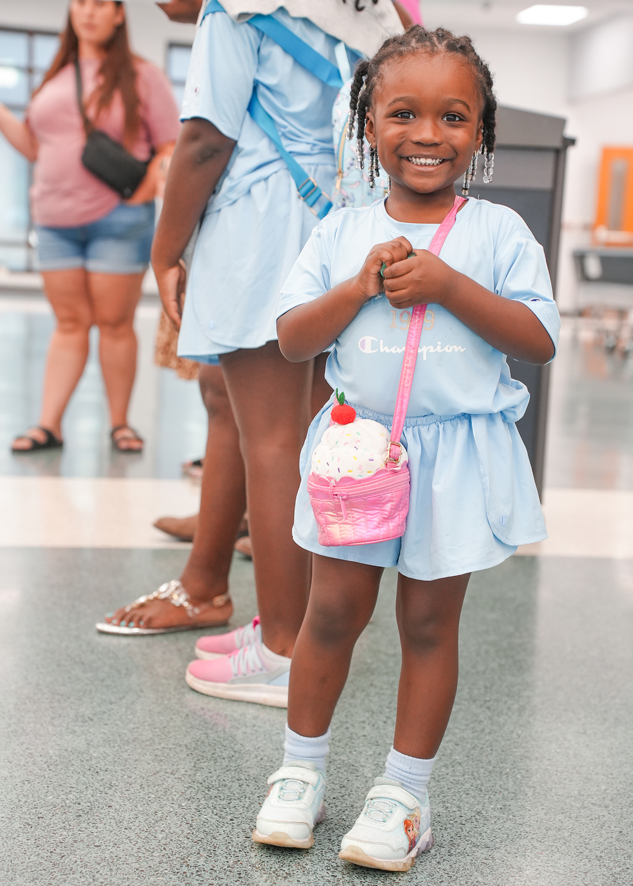 A young girl stands, wearing a blue outfit, and holds a pink purse shaped like a cupcake. Behind her, a few people walk in a hallway.