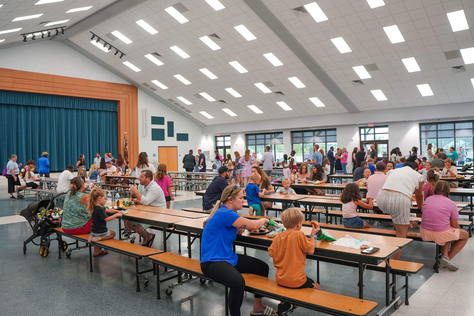 Large dining hall with many people seated at tables. The room is brightly lit with overhead fluorescent lighting.