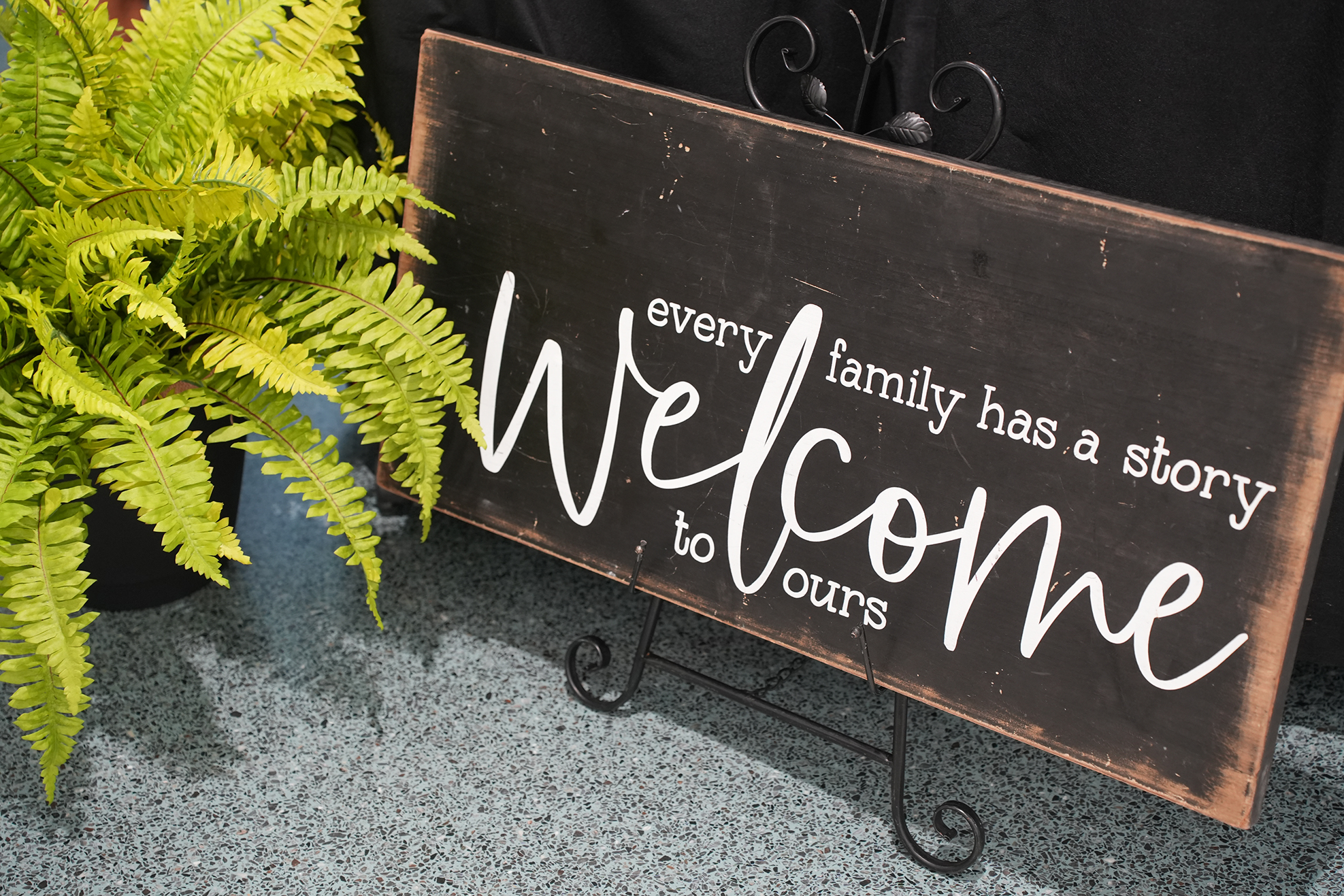 A wooden sign with white lettering reading "welcome every family has a story to ours" stands beside a green fern.