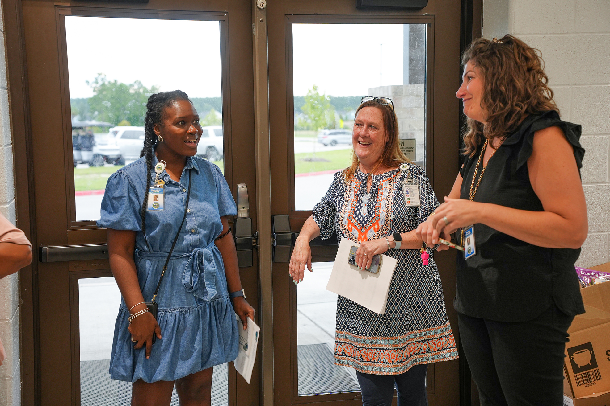 Three women converse near a door; one holds a paper, and another has a box. The background shows an open doorway with vehicles and greenery.