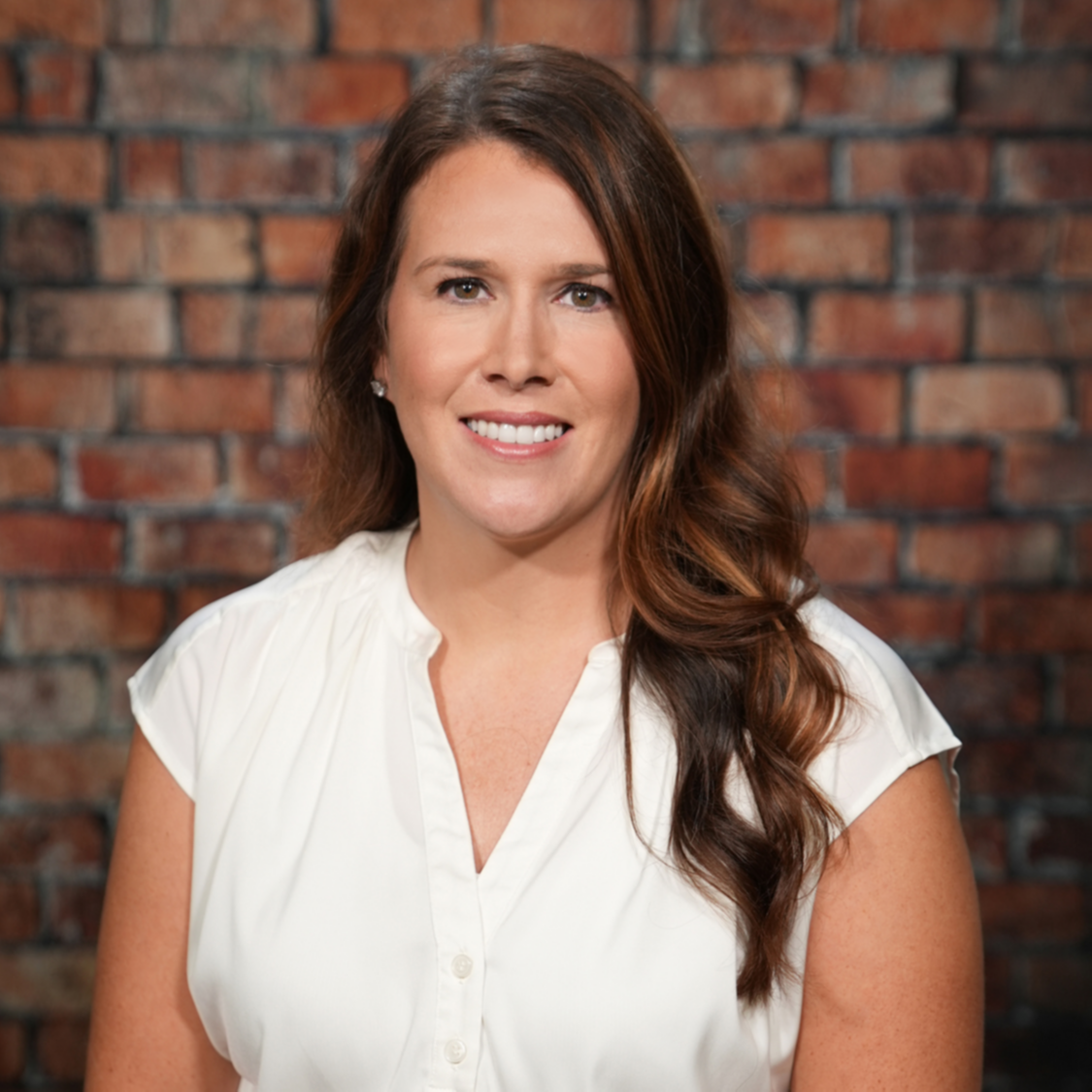 A woman with brown hair smiles at the camera, wearing a white blouse. Behind her is a brick wall.